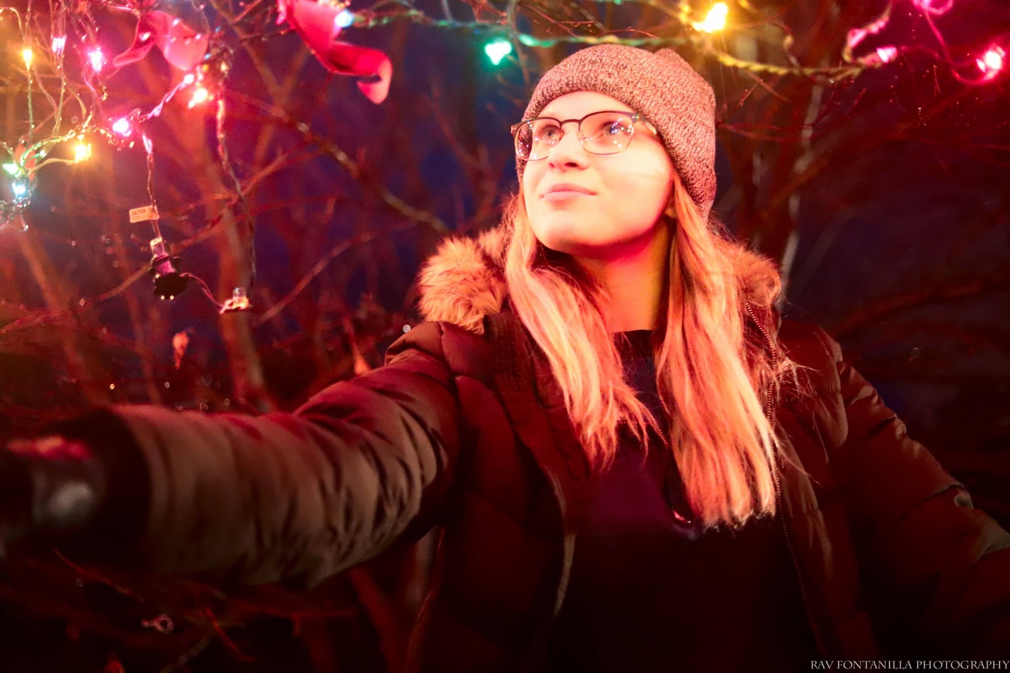 Young woman wearing glasses and a knit hat, taking a selfie outdoors at night with colorful string lights in the background.