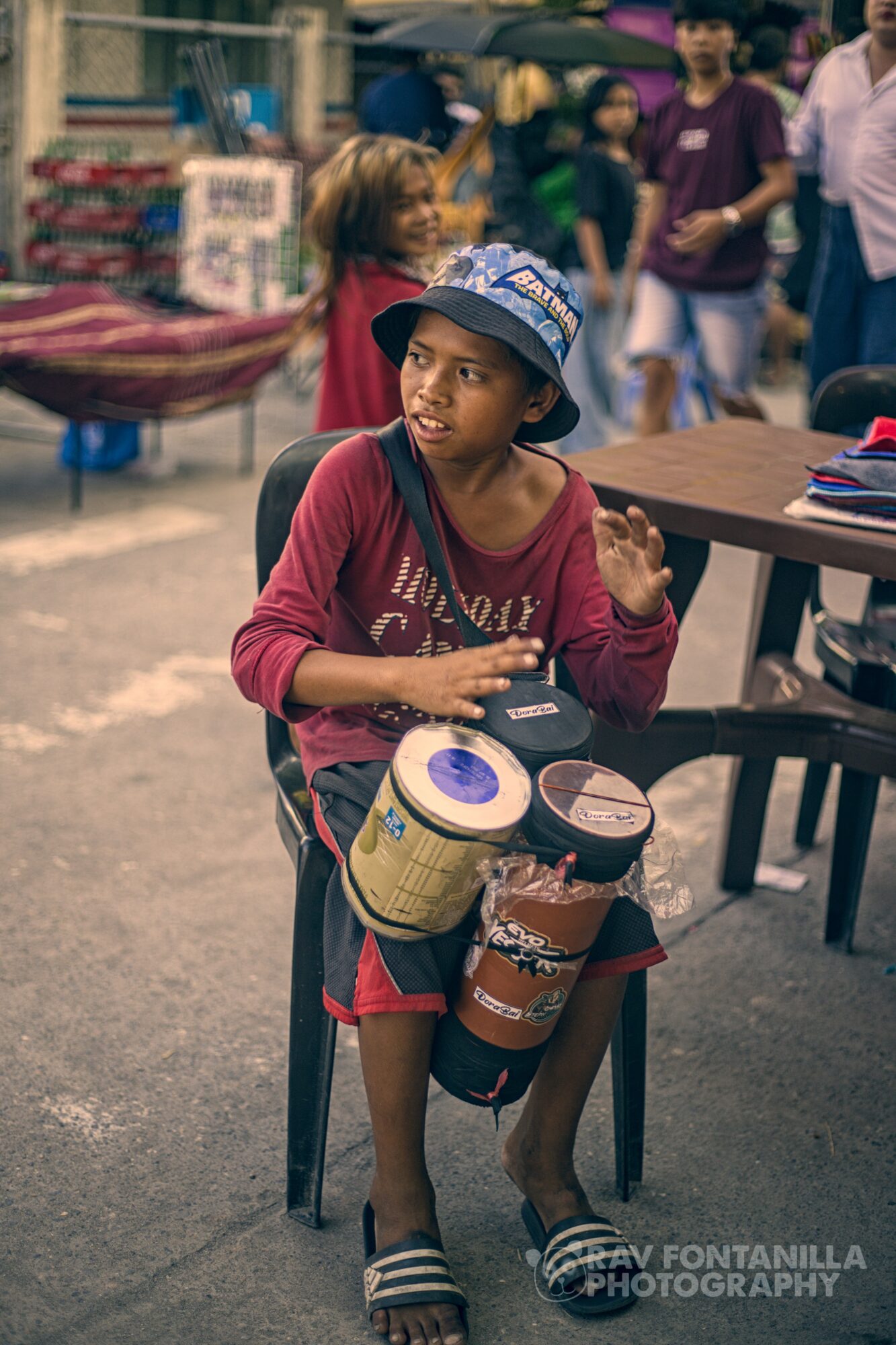 Child sitting on a chair with drums and a hat, surrounded by people at an outdoor market or event.