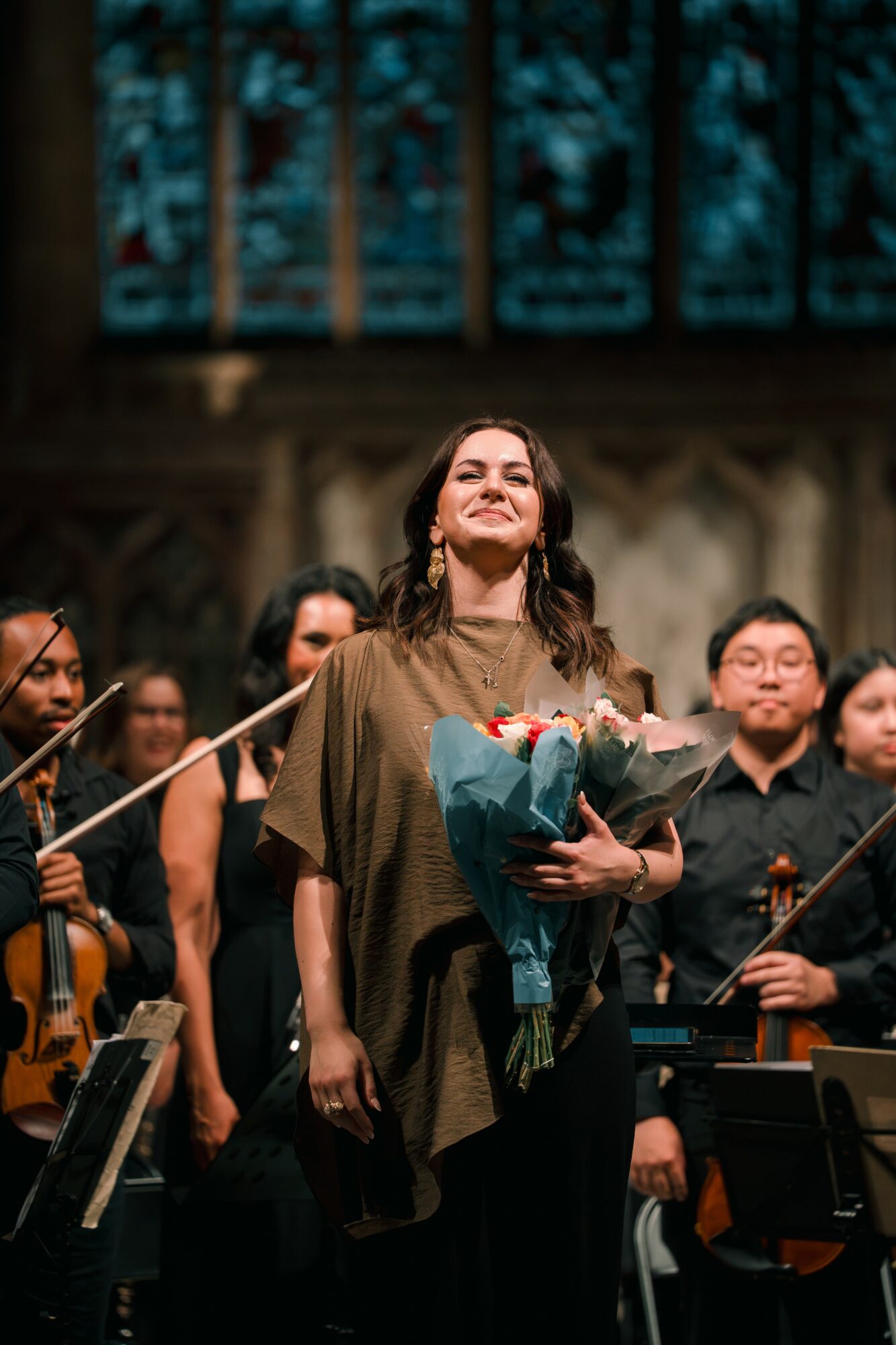 Woman holding a bouquet of flowers on stage with orchestra behind her, smiling, in a concert hall.