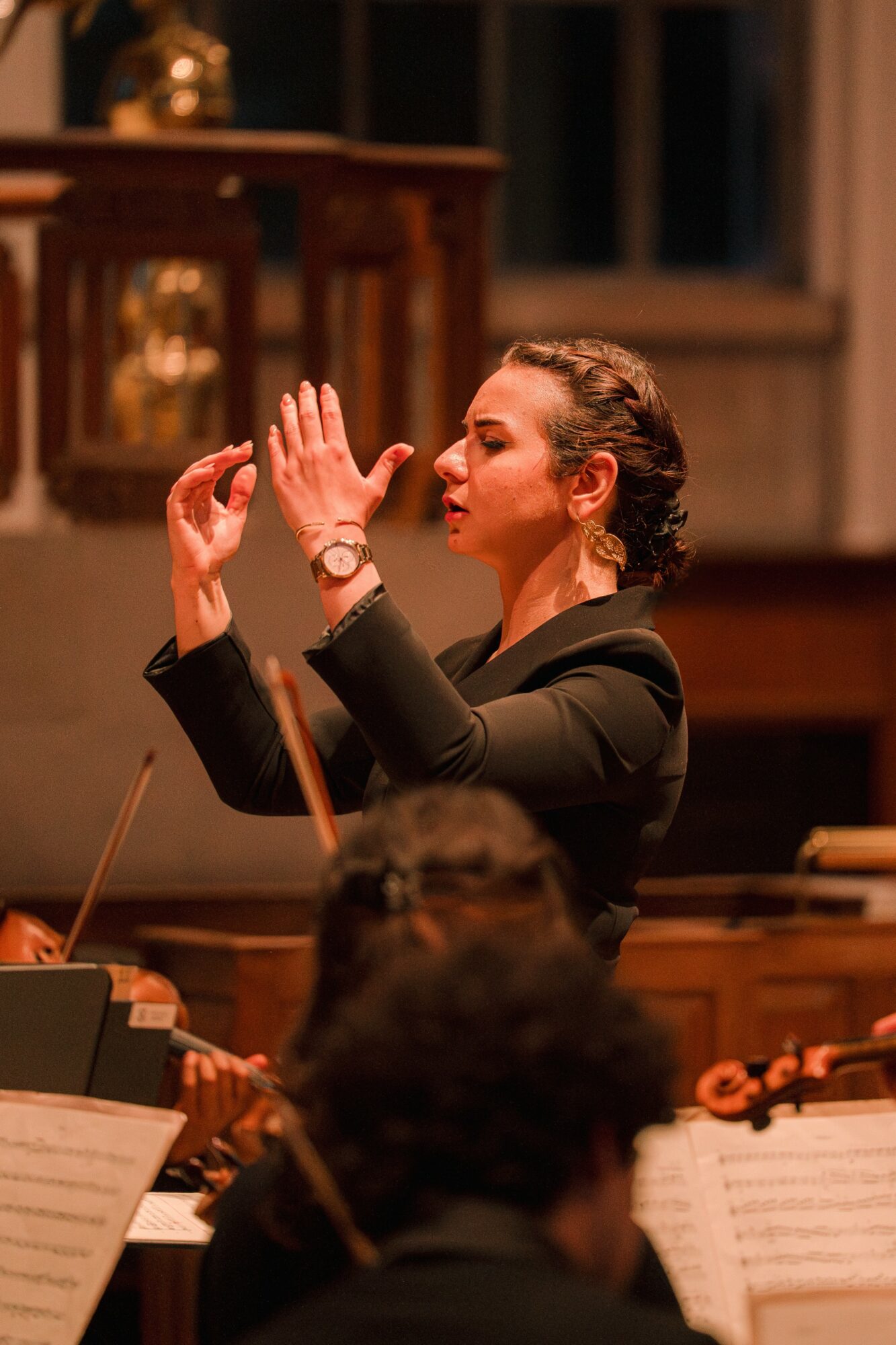Woman conducting an orchestra, raising her hands, in a formal setting with music stands and sheet music.