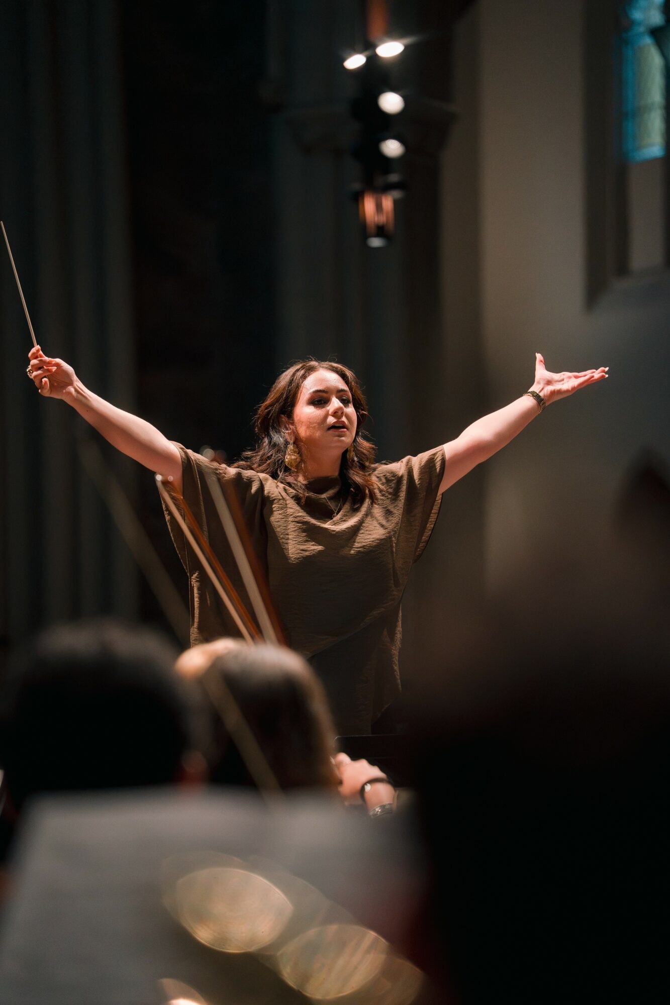 Woman on stage with arms outstretched, holding a baton, performing in a theater setting with stage lights and curtains.
