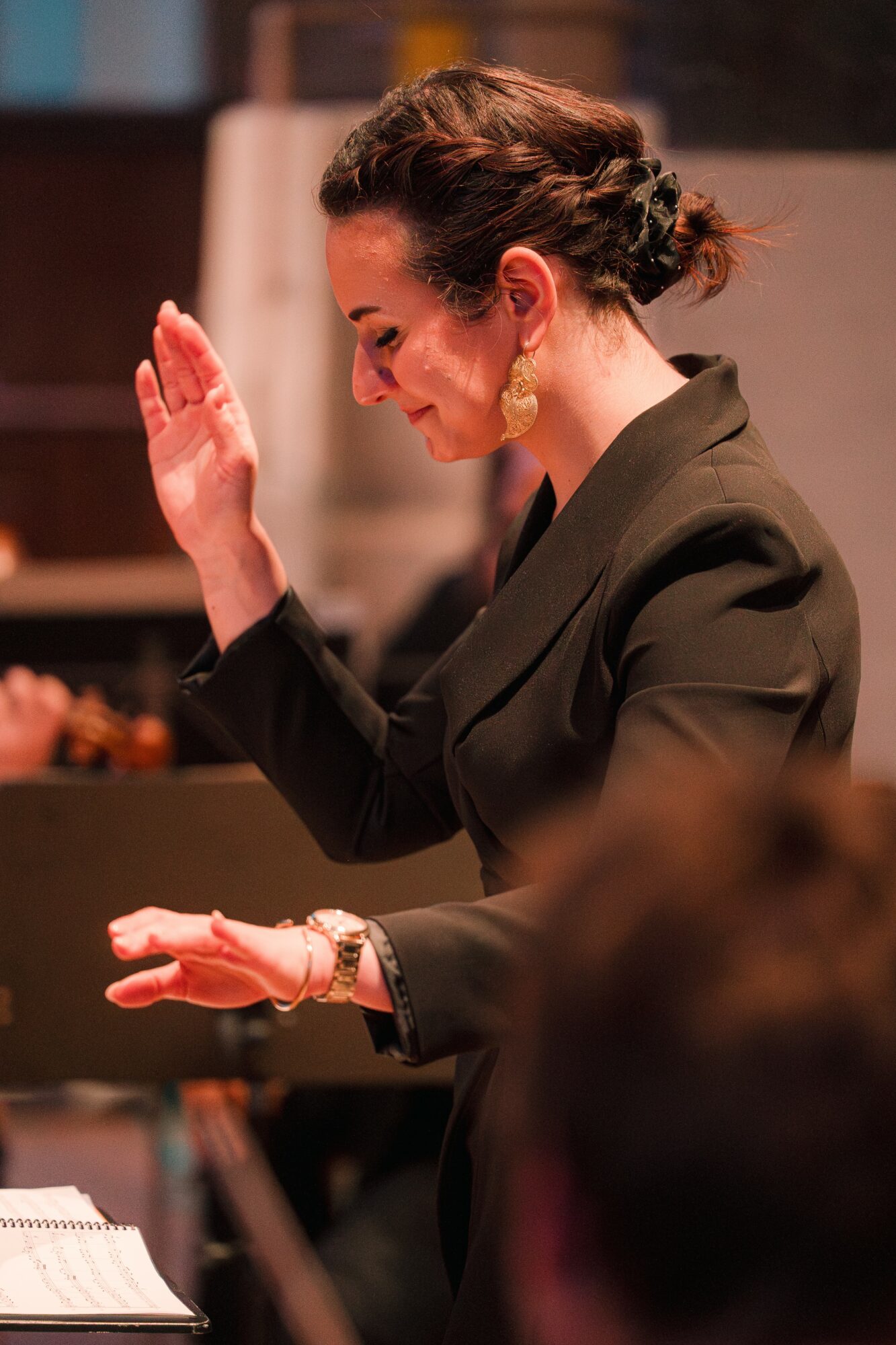 Woman with dark hair in an updo, wearing a dark blazer, raising her right hand and looking down, in an indoor setting.