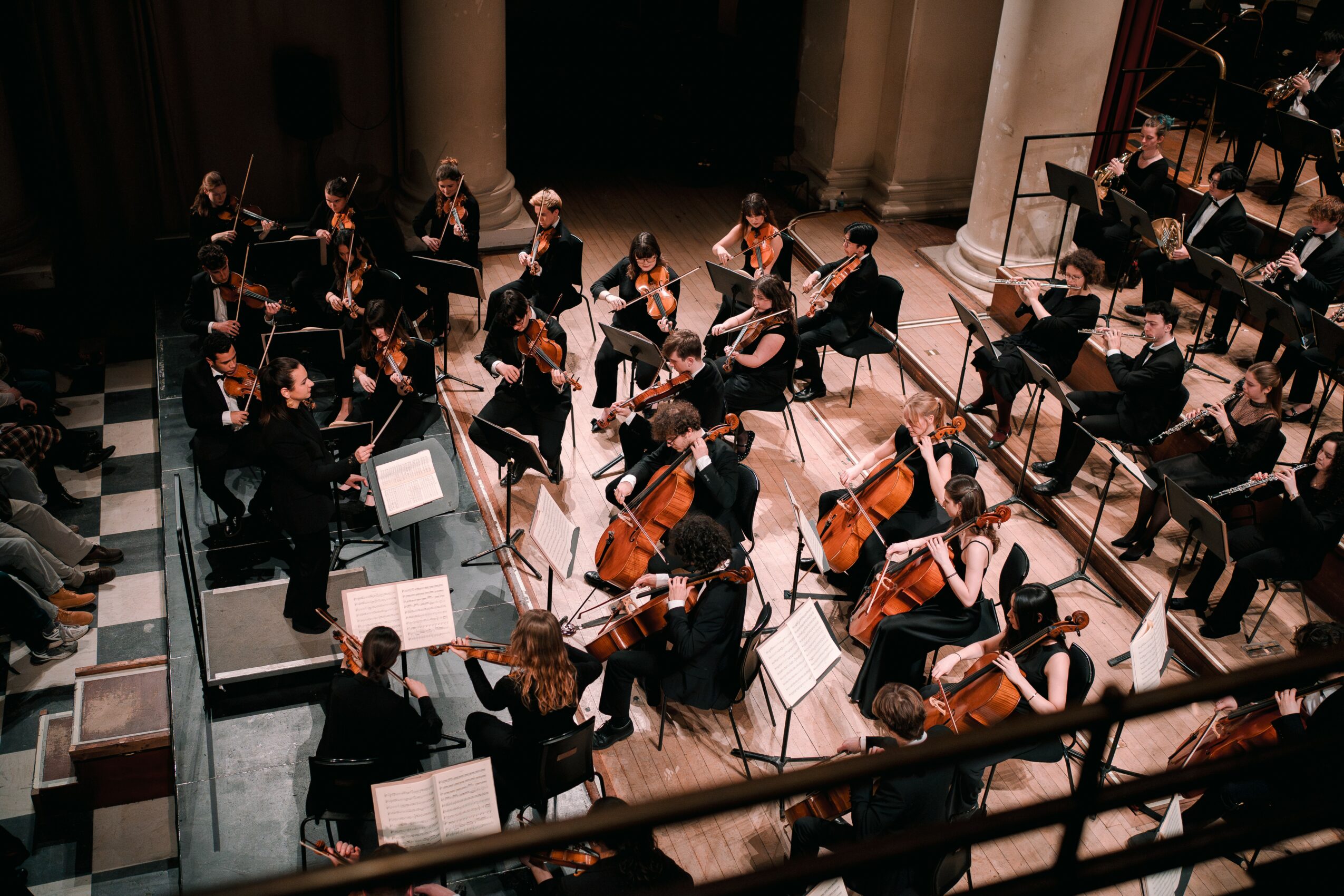 Orchestra performing on stage with musicians playing string, woodwind, and percussion instruments, viewed from above.
