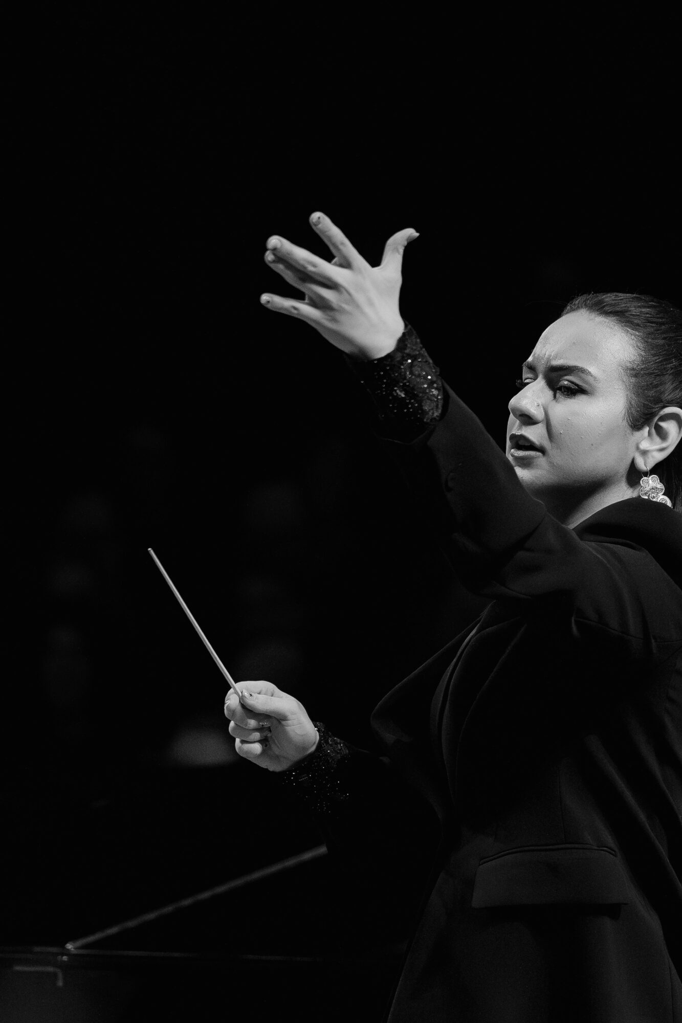Conductor leading an orchestra with raised hand and baton, focused expression, black background.