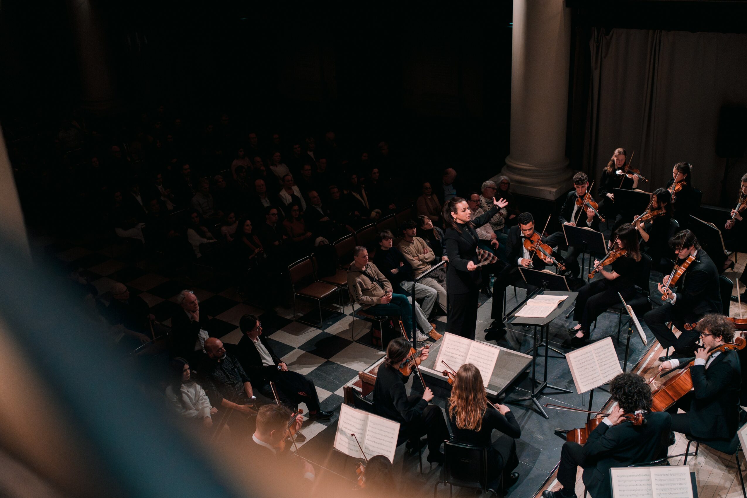 Orchestra performing on stage with musicians reading sheet music, audience watching, in a dimly lit concert hall.