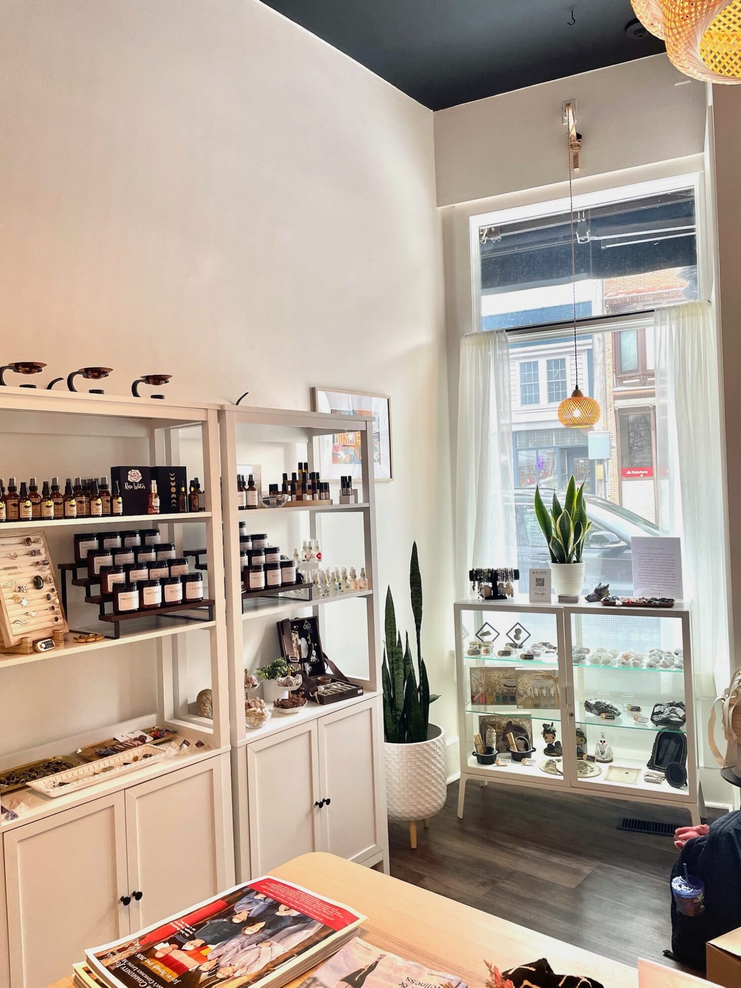 Interior of a shop with shelves of bottles and boxes, a window with a plant, and a display case, sunlight streaming in.