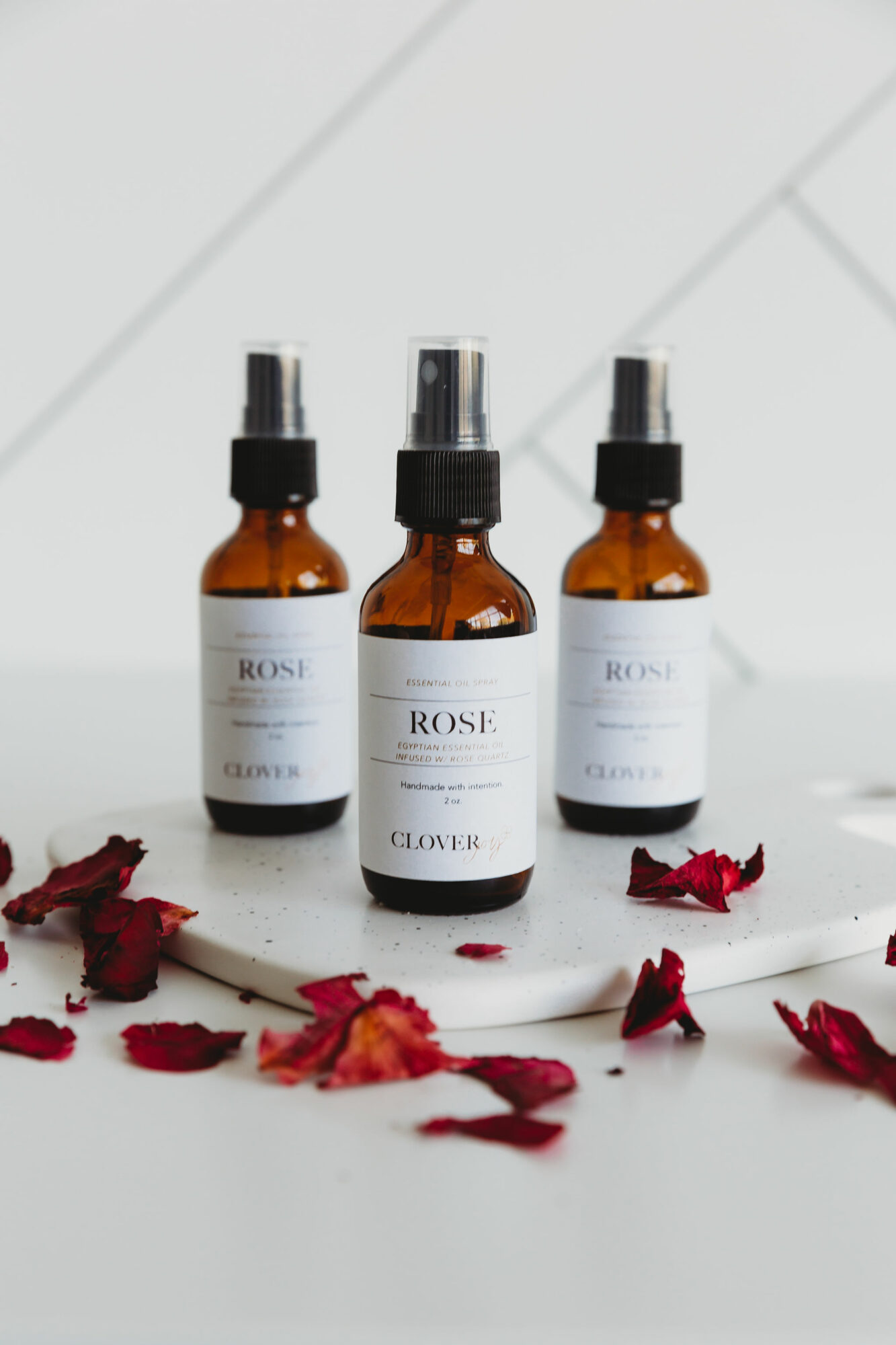 Three amber spray bottles labeled 'Rose' and 'Clover' on a white surface with scattered red flower petals.