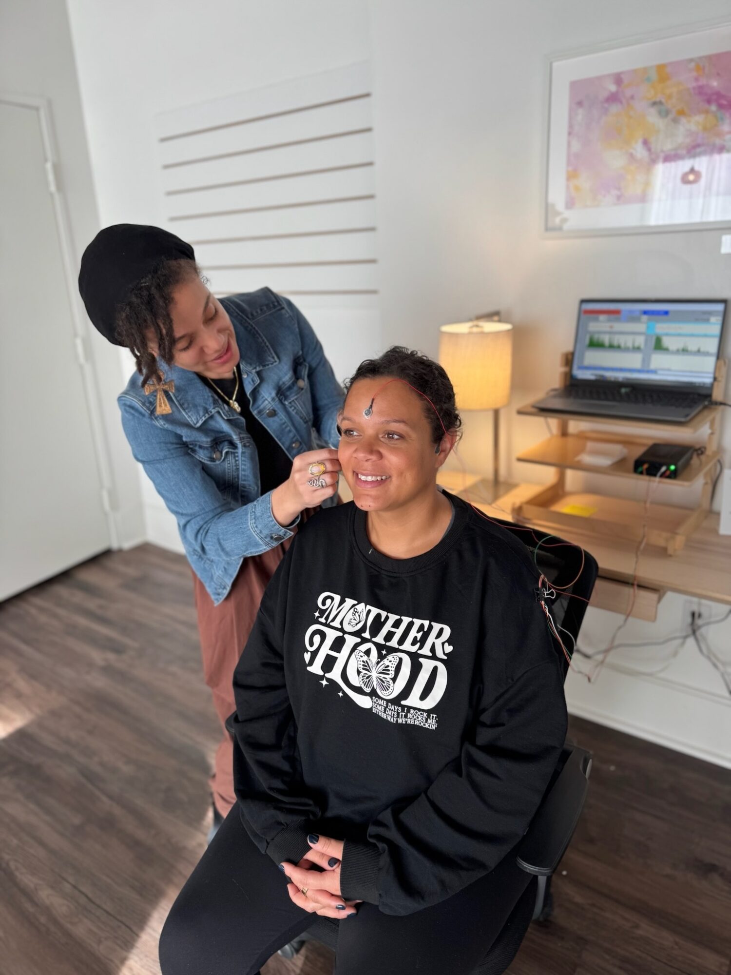 Person applying makeup to smiling woman seated in a room with a desk, computer, and framed artwork on the wall.
