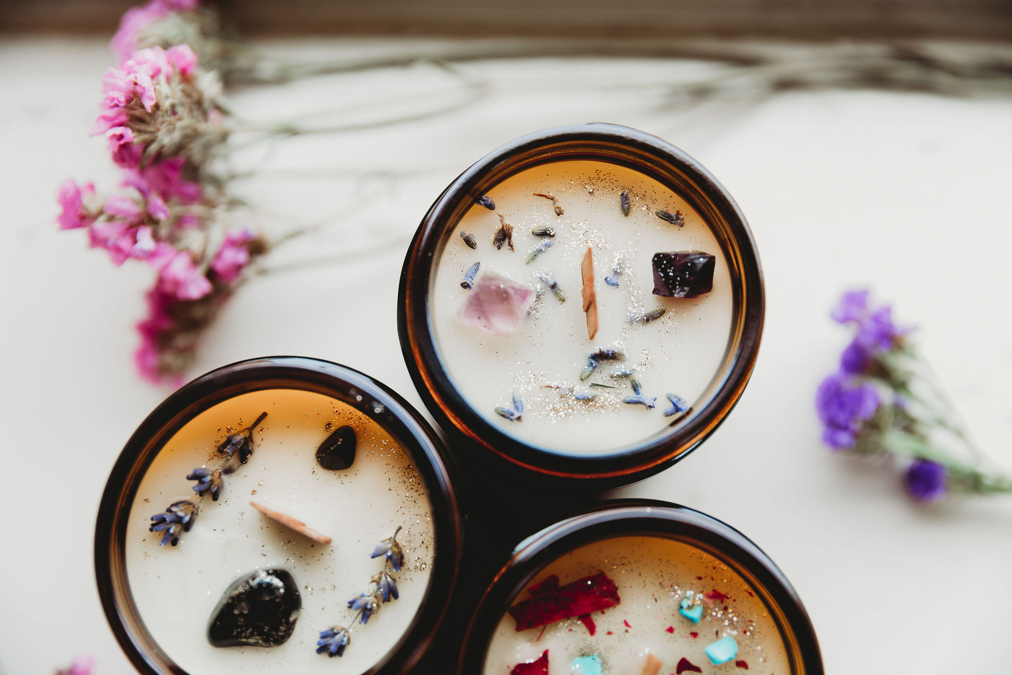 Three candles with dried flowers and herbs in glass containers on a white surface, with pink and purple flowers nearby.