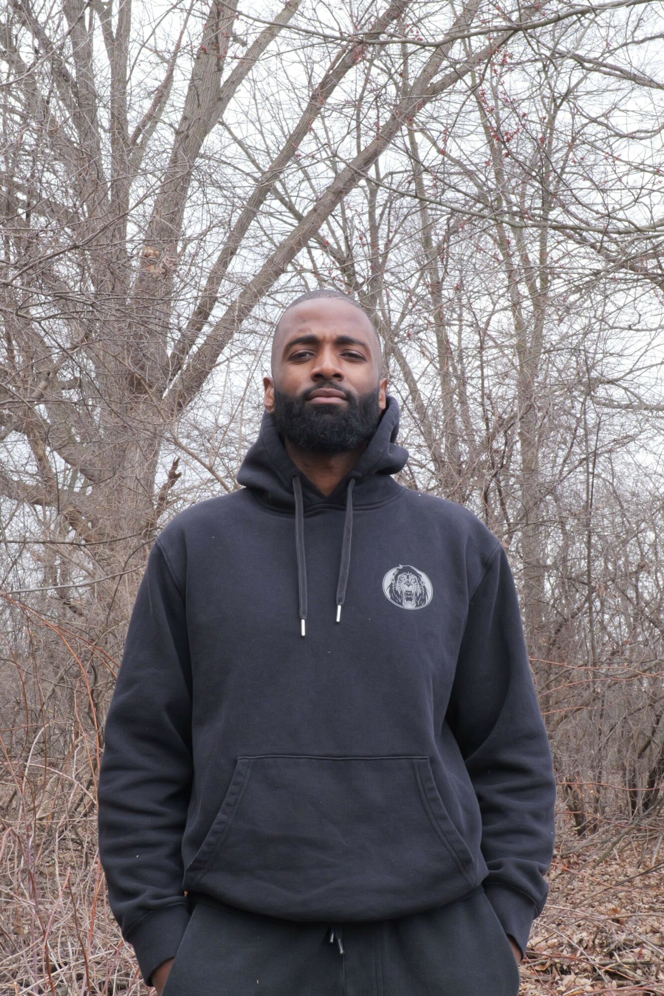 Man standing outdoors among leafless trees, wearing a black hoodie with a logo, looking at the camera.