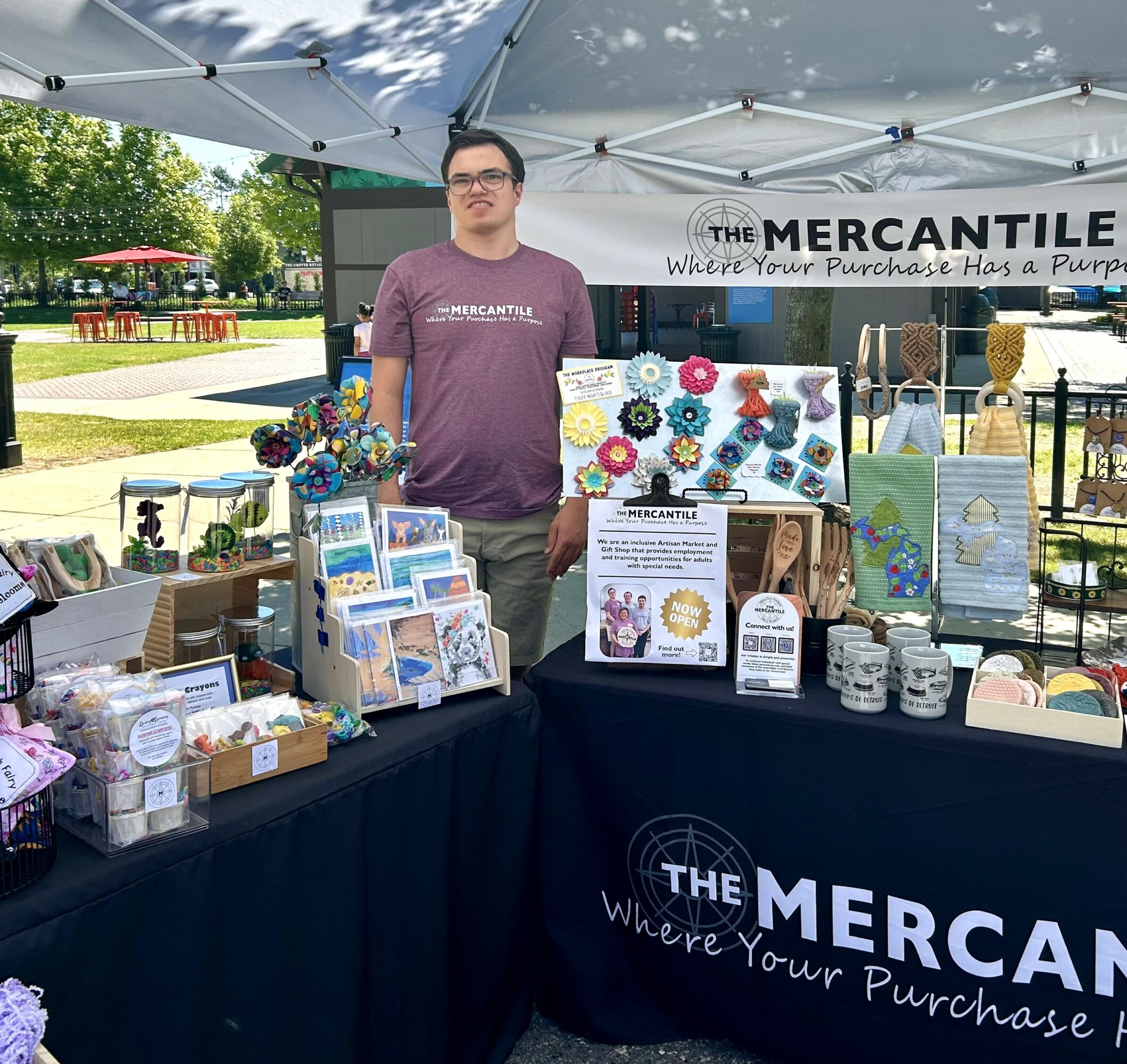 Person standing behind a booth with colorful crafts and signs, under a tent with a banner reading 'The Mercantile'.