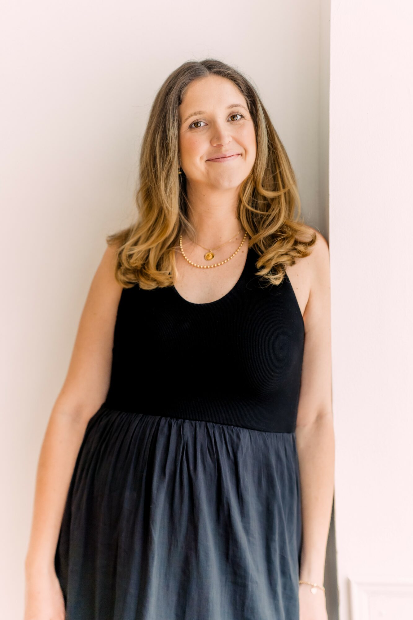 Young woman with shoulder-length wavy hair smiling, wearing a black sleeveless top and a dark skirt, standing indoors.