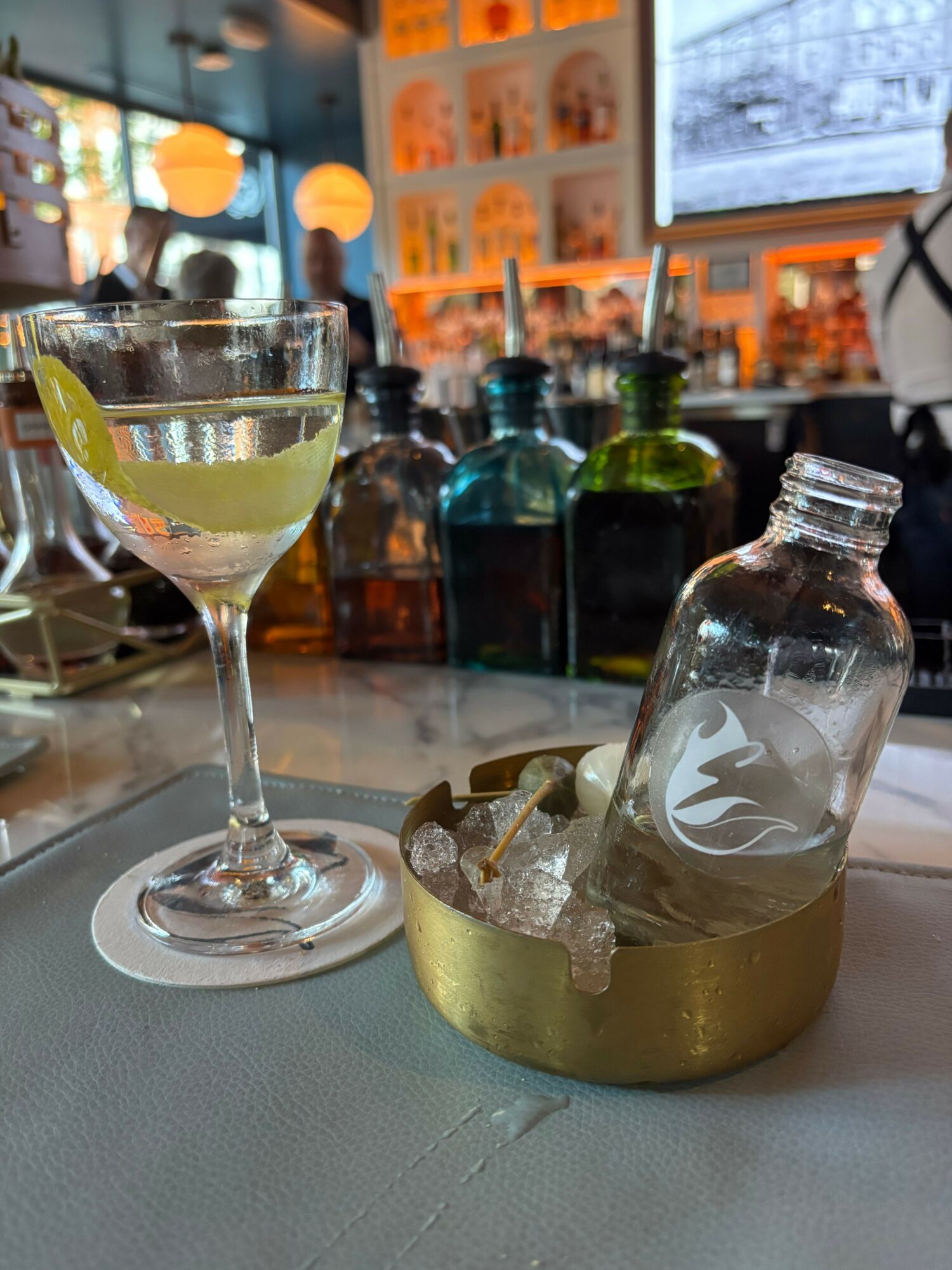 A cocktail glass with a lemon wedge, a bottle with a flame logo, and various bottles in the background.