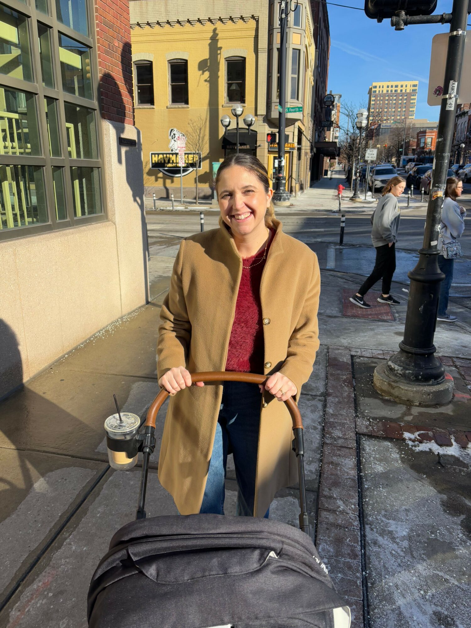 Woman in beige coat pushing a stroller on city sidewalk, buildings and street scene in background.