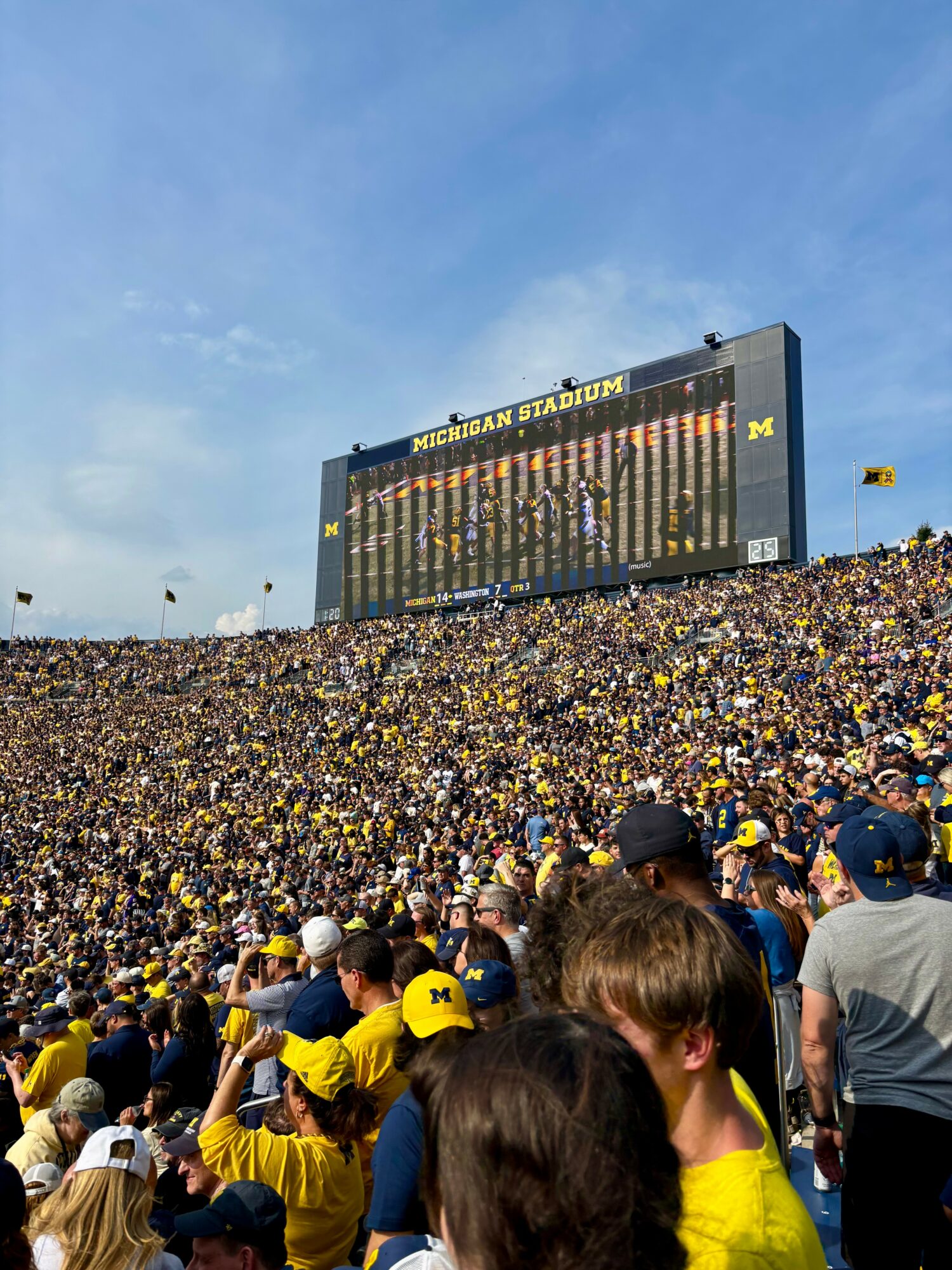 Crowd of people wearing yellow and blue at Michigan Stadium during daytime, with a large scoreboard displaying game information.
