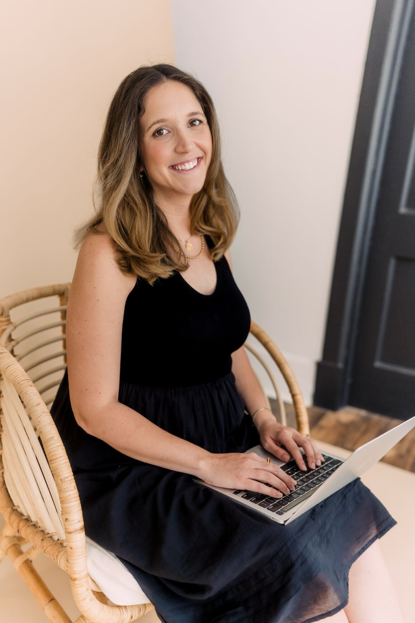 Young woman with shoulder-length hair sitting on a wicker chair, smiling, using a laptop, in a room with beige and gray walls.