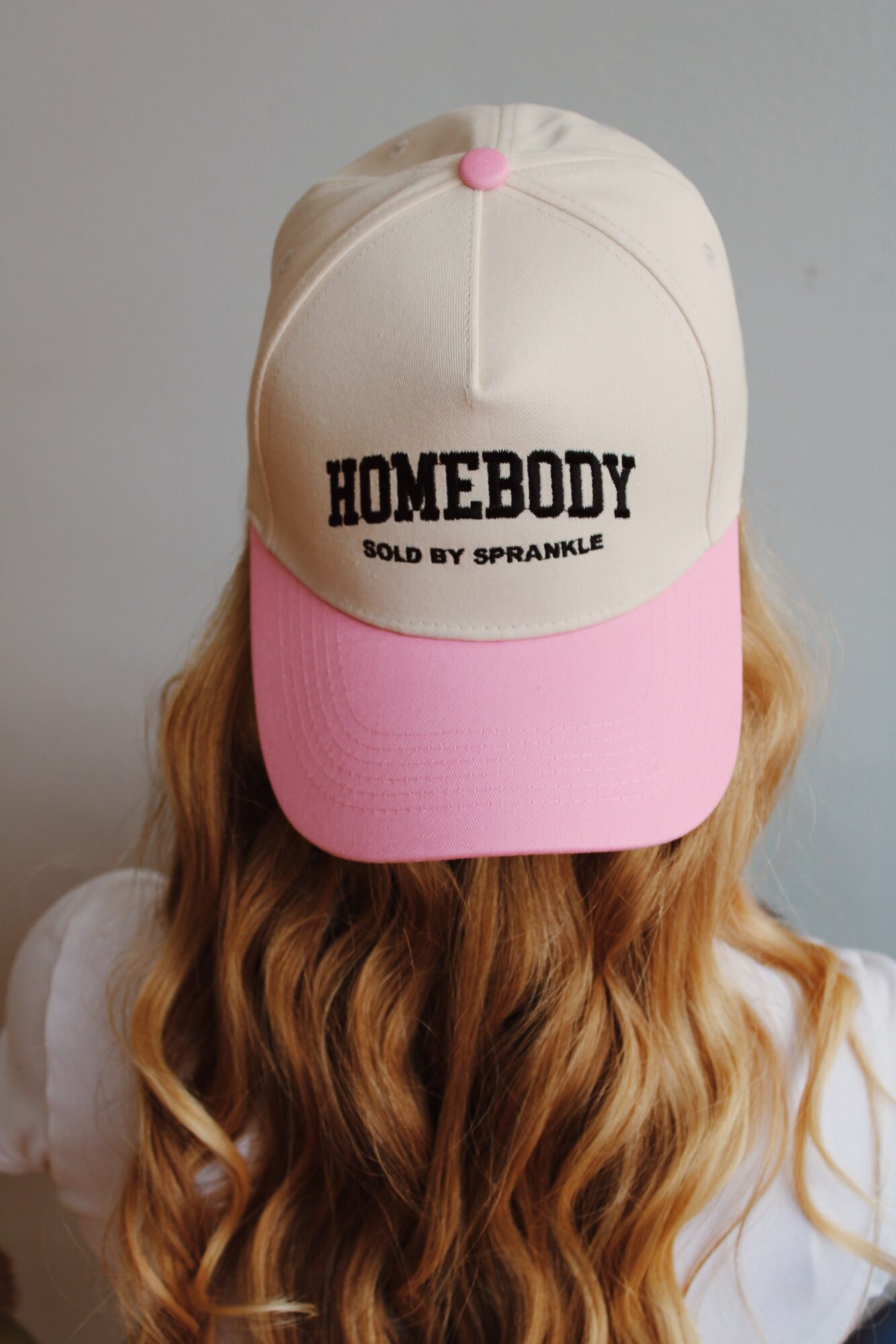 Person with long wavy hair wearing a beige and pink cap with text, facing away from the camera.