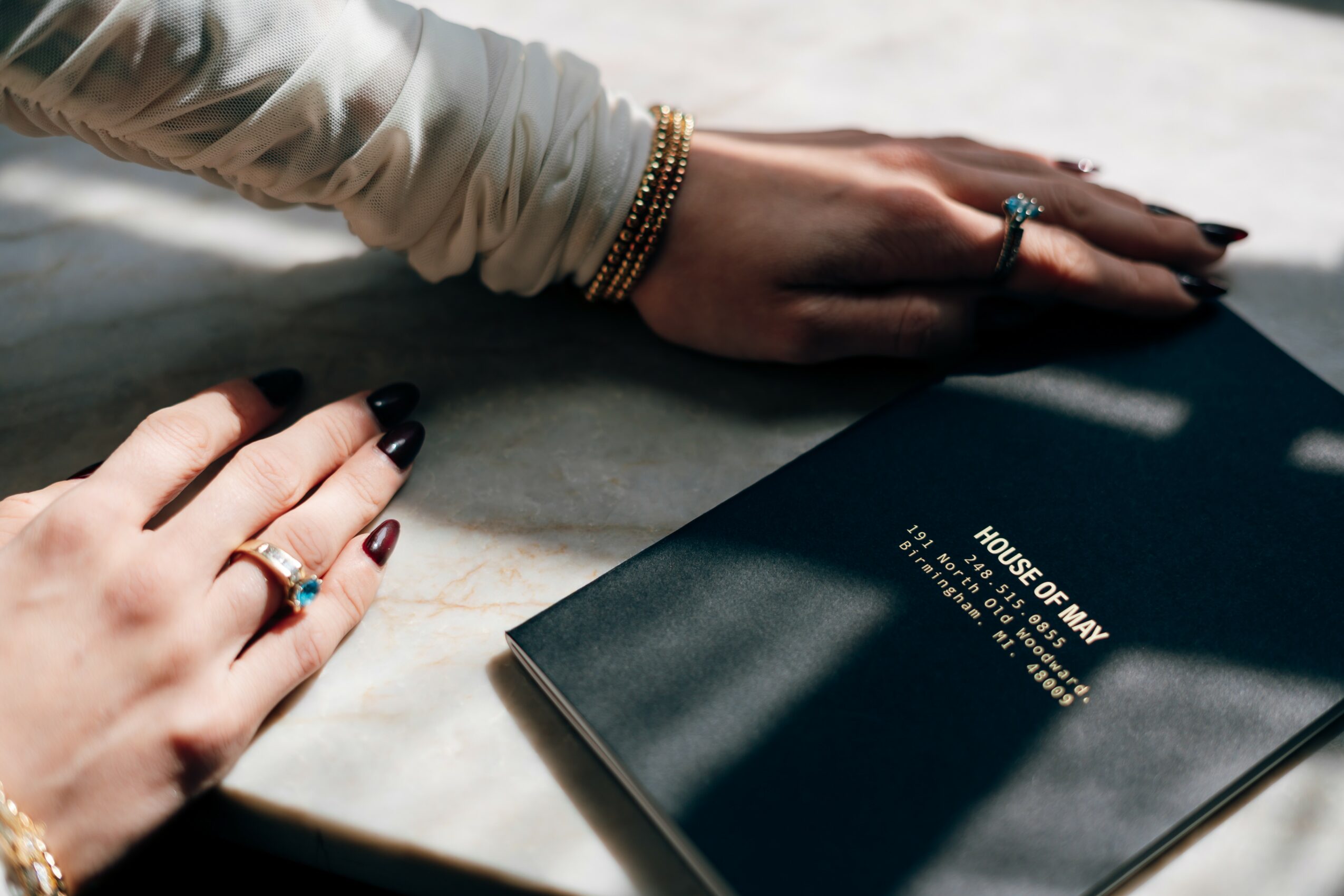 Two hands with rings and bracelets rest on a marble surface near a dark blue book titled 'HOUSE OF MAY'.