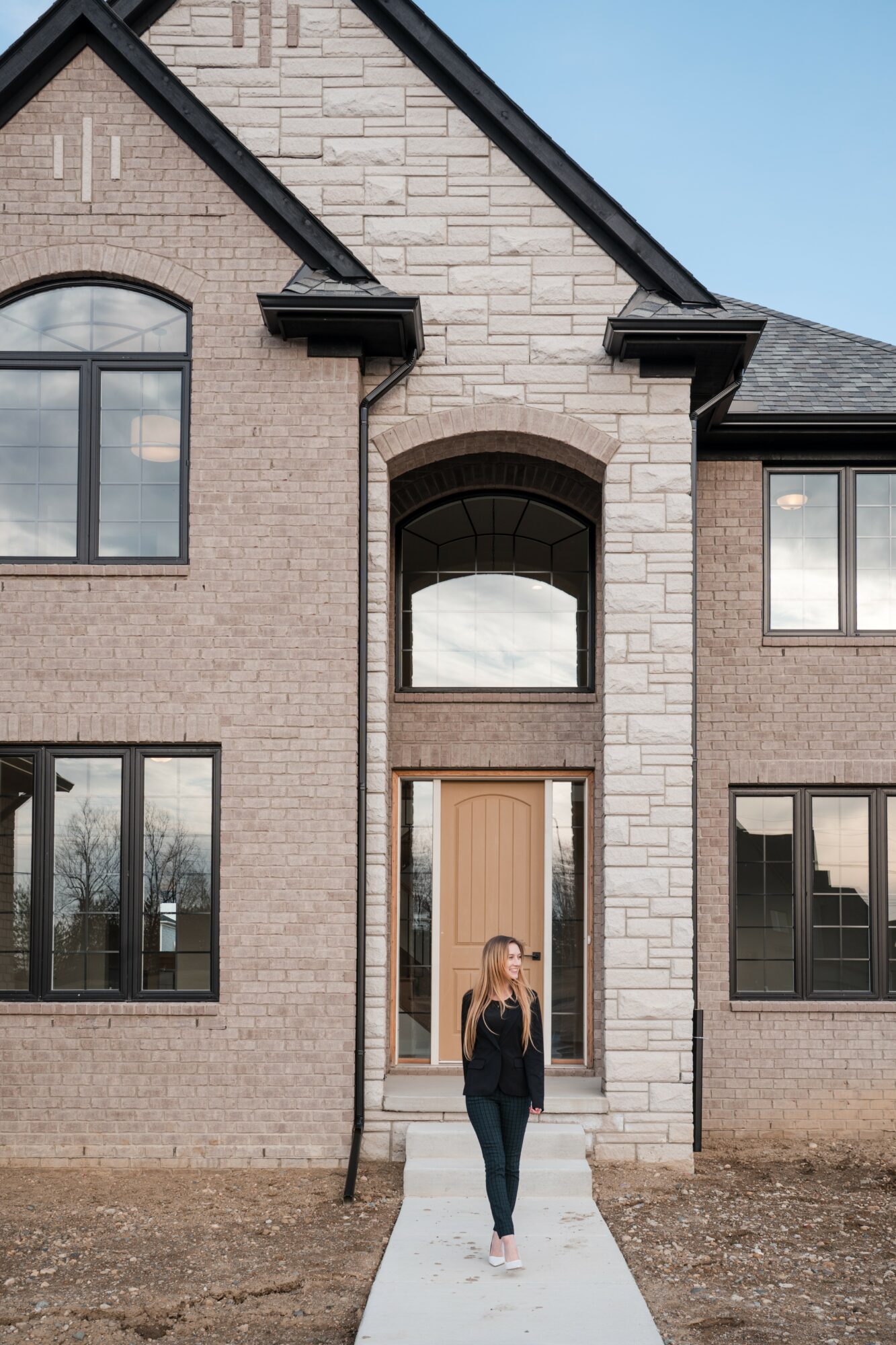 Front view of a brick house with arched window and entrance, woman walking on sidewalk, clear sky in background.