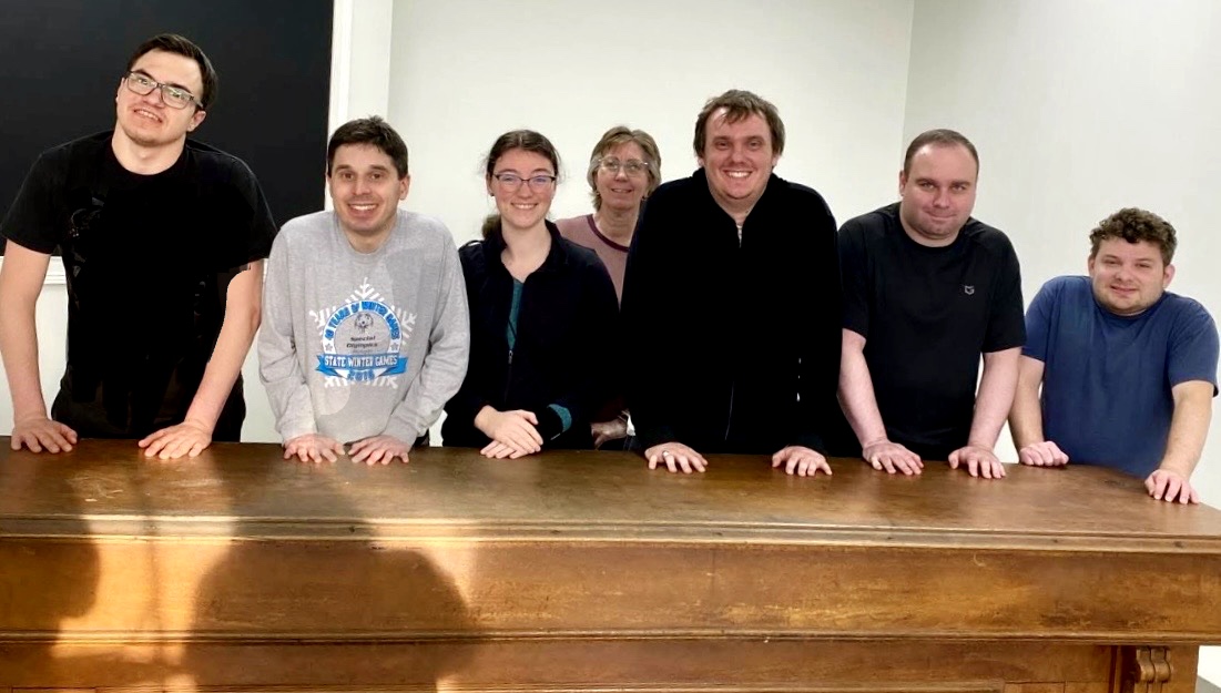 Group of seven people standing behind a wooden table, smiling, in a room with plain white walls.