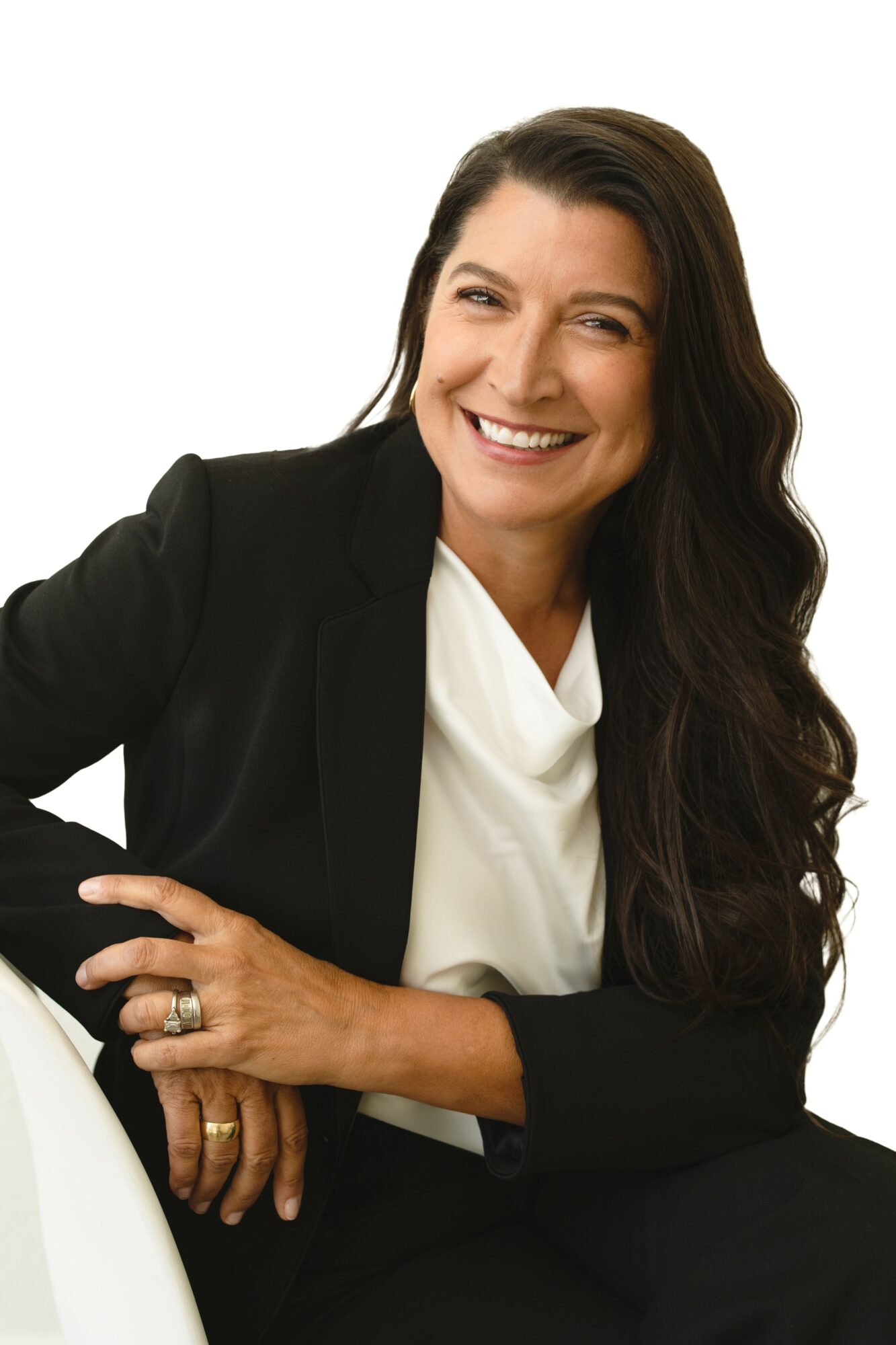 Smiling woman with long dark hair, wearing a black blazer and white blouse, sitting with her hand on her knee.