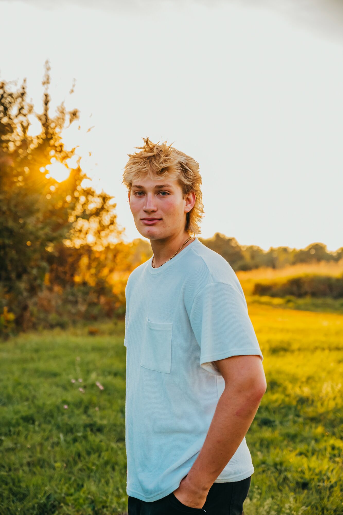 Young person with blond hair standing outdoors in a grassy field during sunset, wearing a light blue T-shirt.