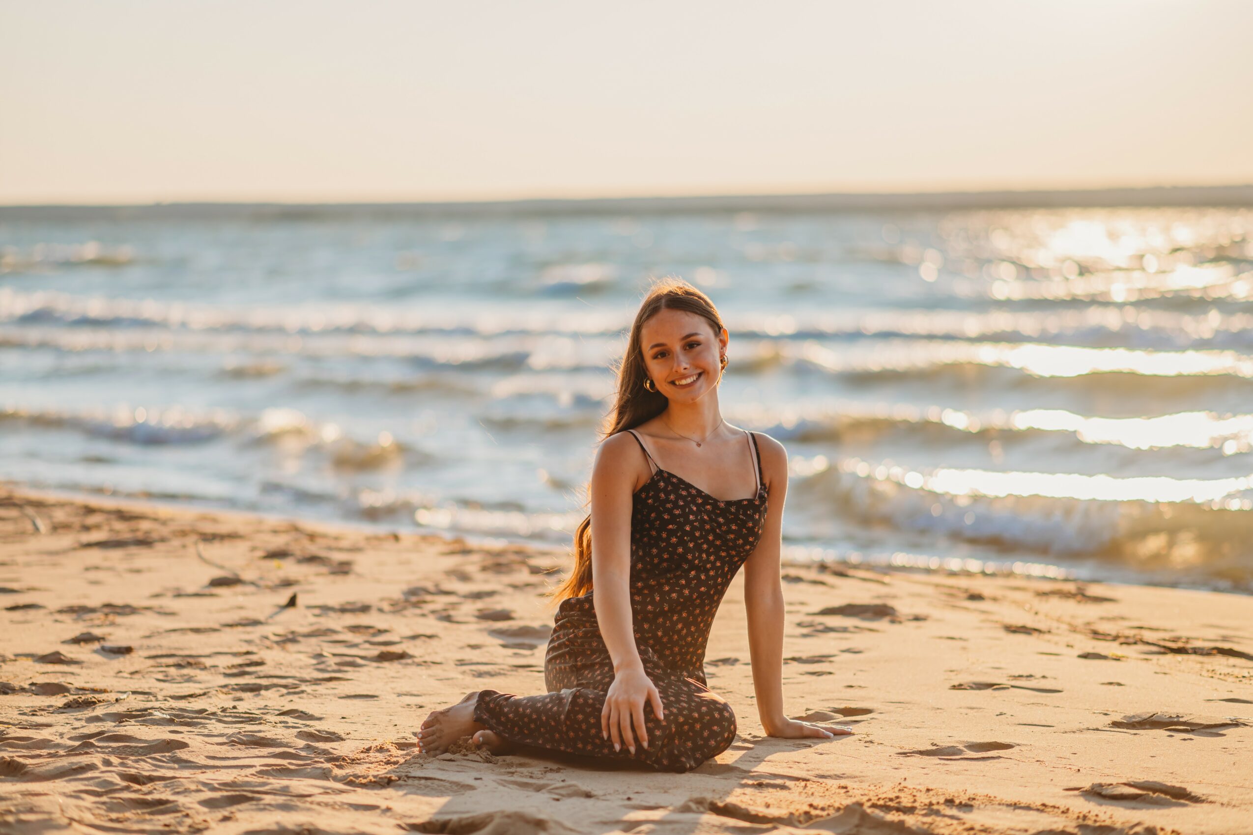 Young woman sitting on sandy beach with ocean waves in background, smiling, wearing a polka dot dress, during sunset.