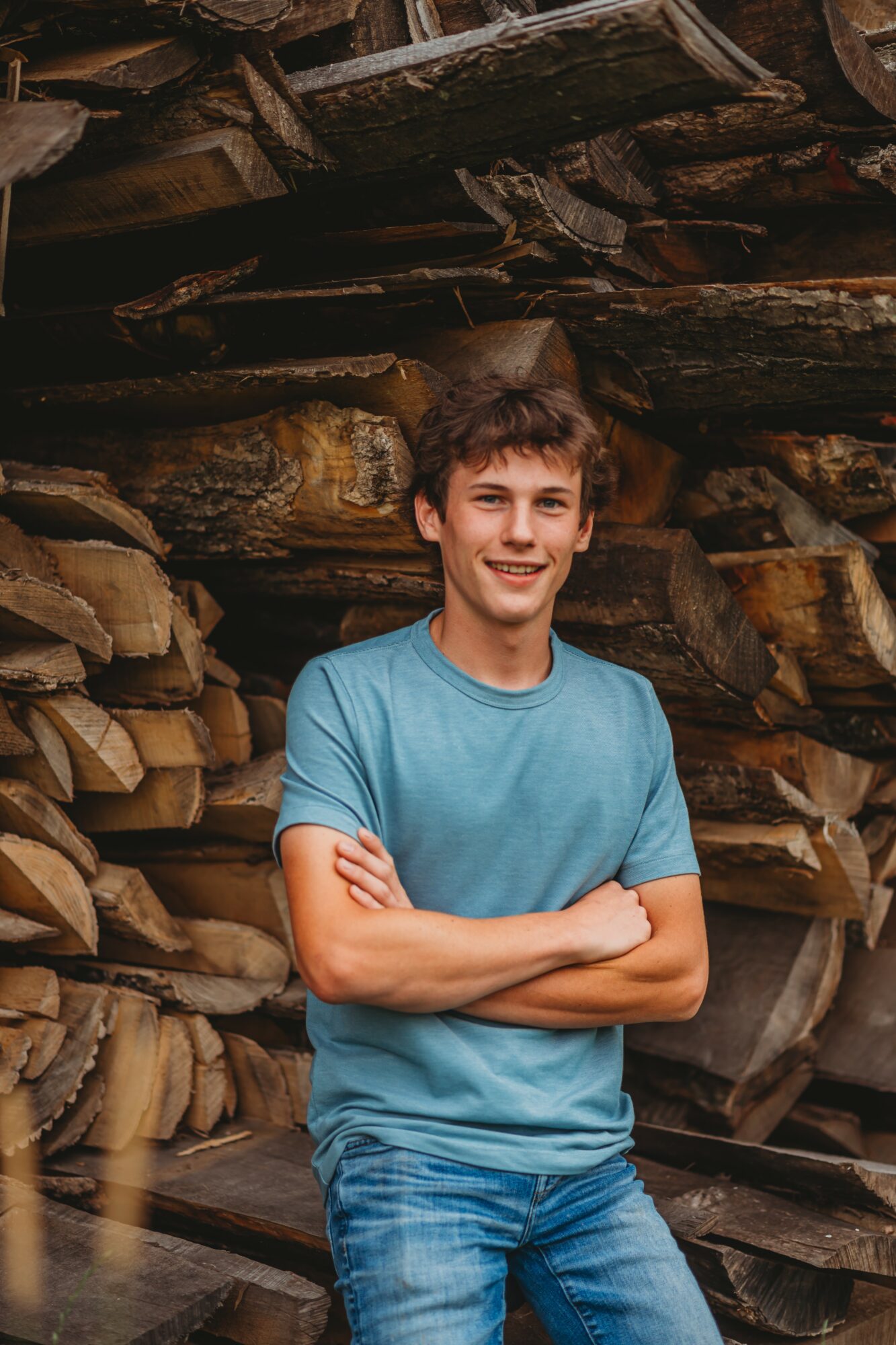 Young man with short brown hair smiling, wearing a blue t-shirt and jeans, standing in front of stacked logs.