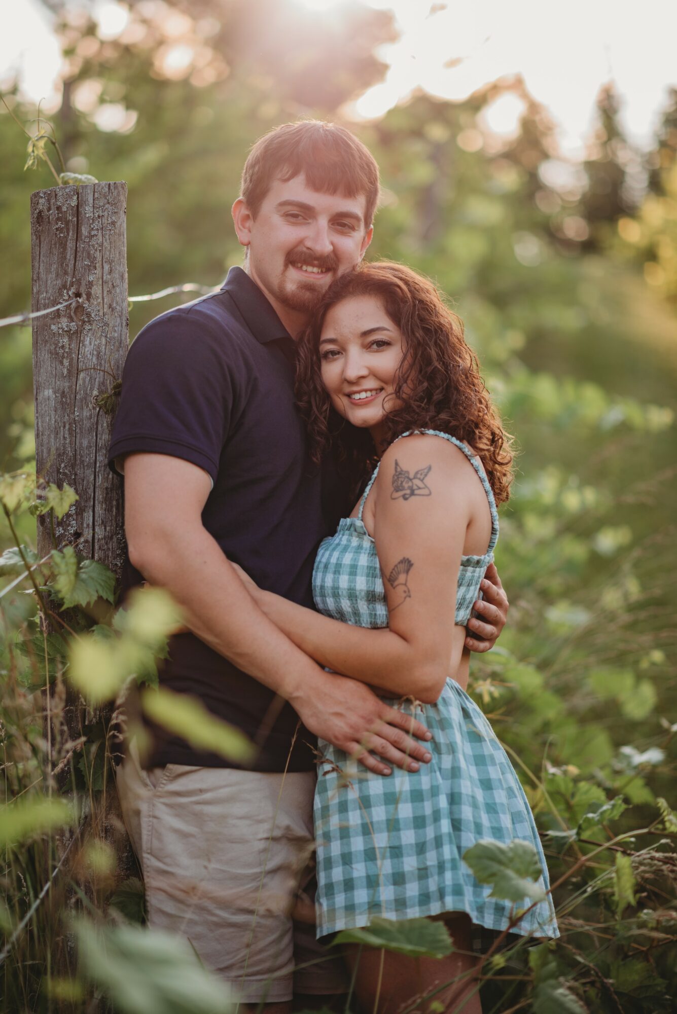 A smiling couple stands outdoors, embracing each other, with trees and sunlight in the background.