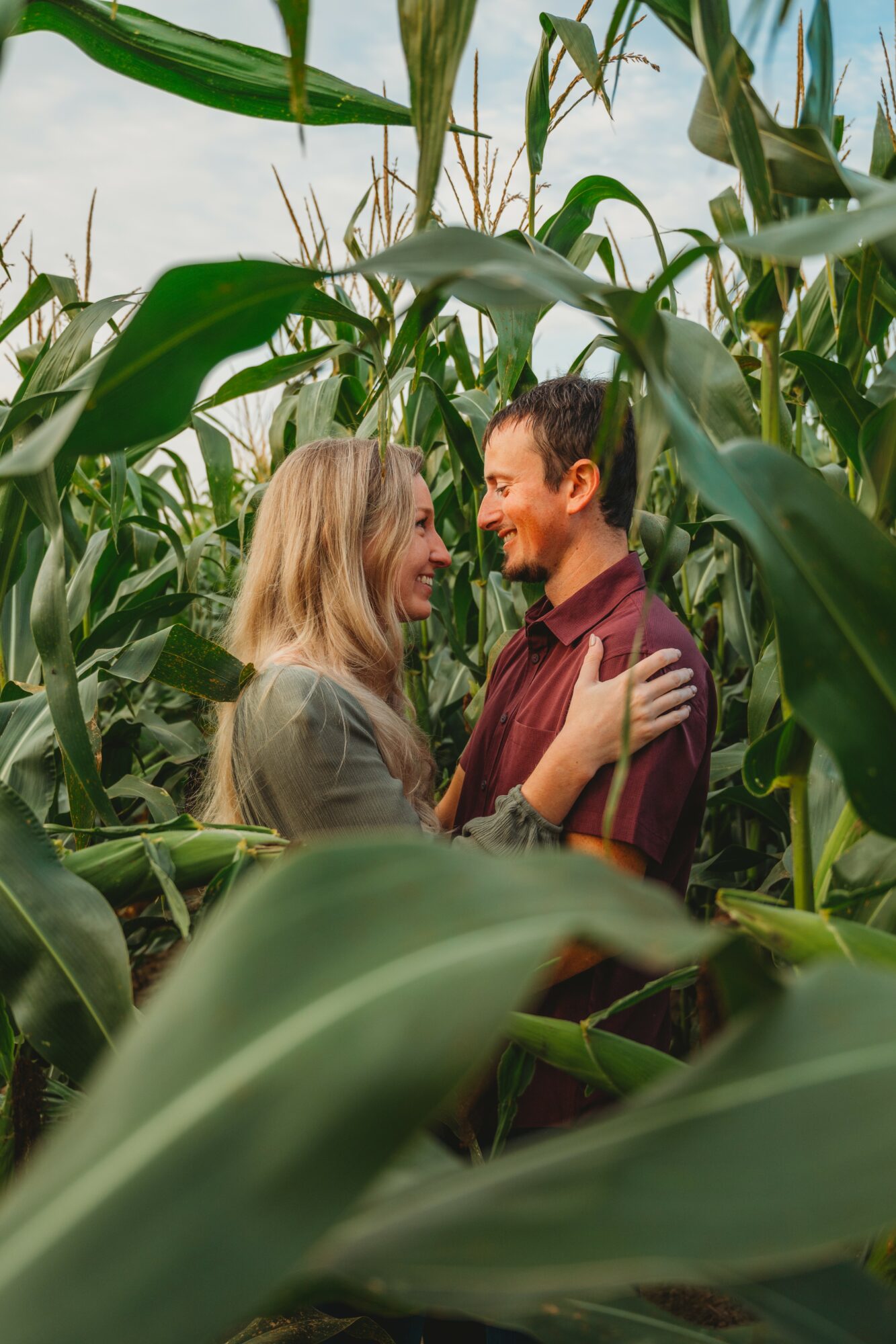 A couple stands close together among tall green corn plants, smiling and looking at each other.