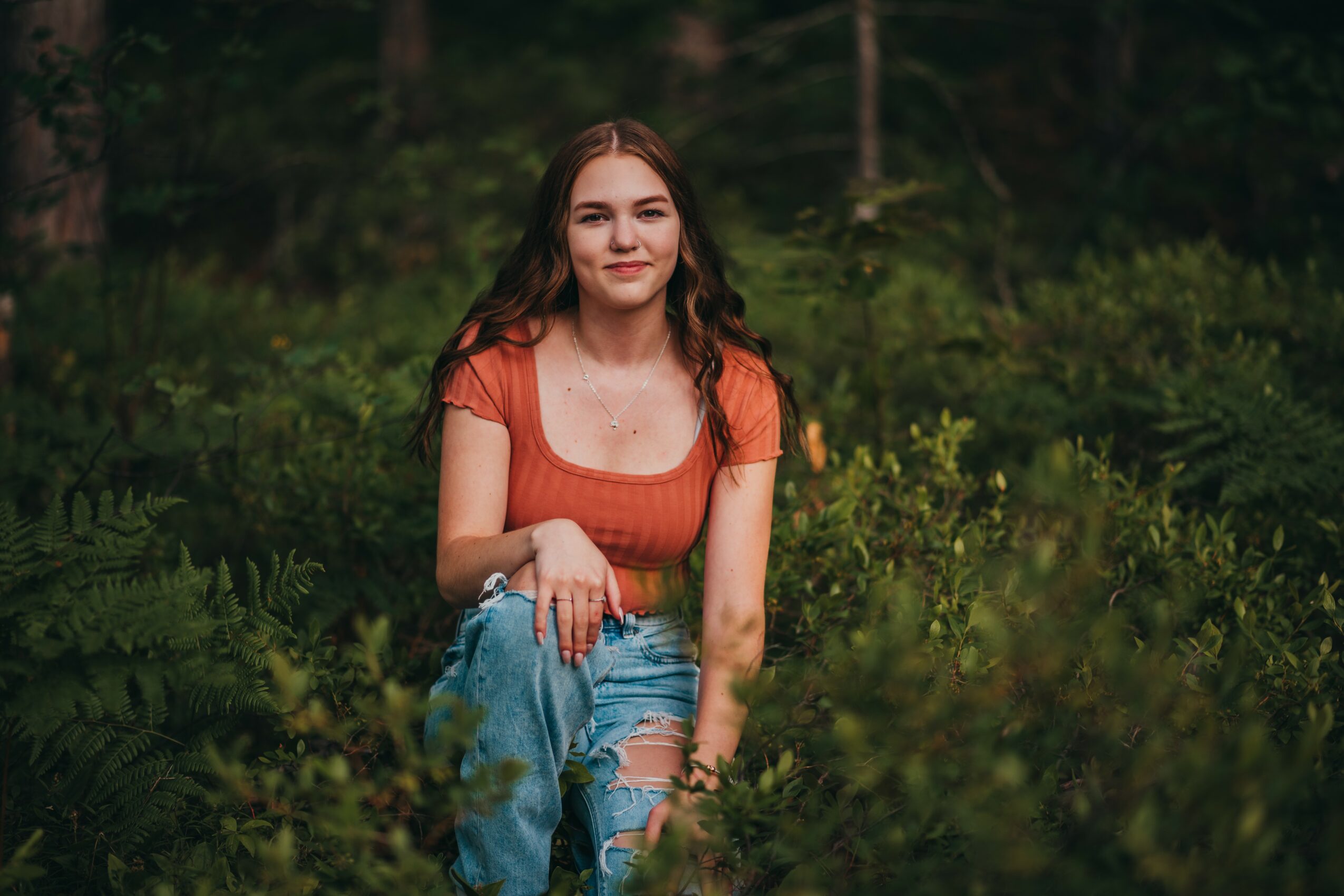 Young woman with long hair sitting outdoors among greenery, smiling, wearing an orange top and ripped jeans.