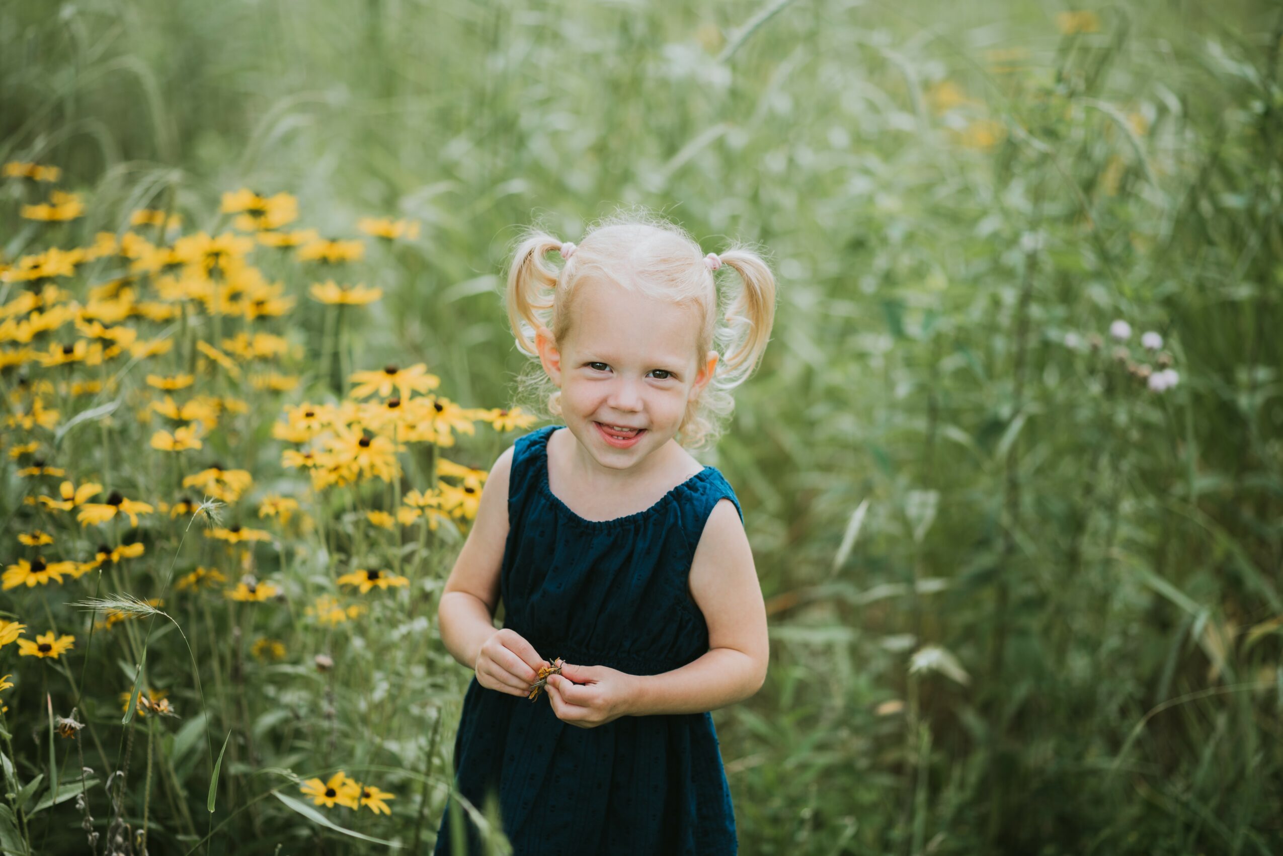 Young girl with blonde hair in pigtails, wearing a dark blue dress, standing outdoors among yellow flowers and green foliage.
