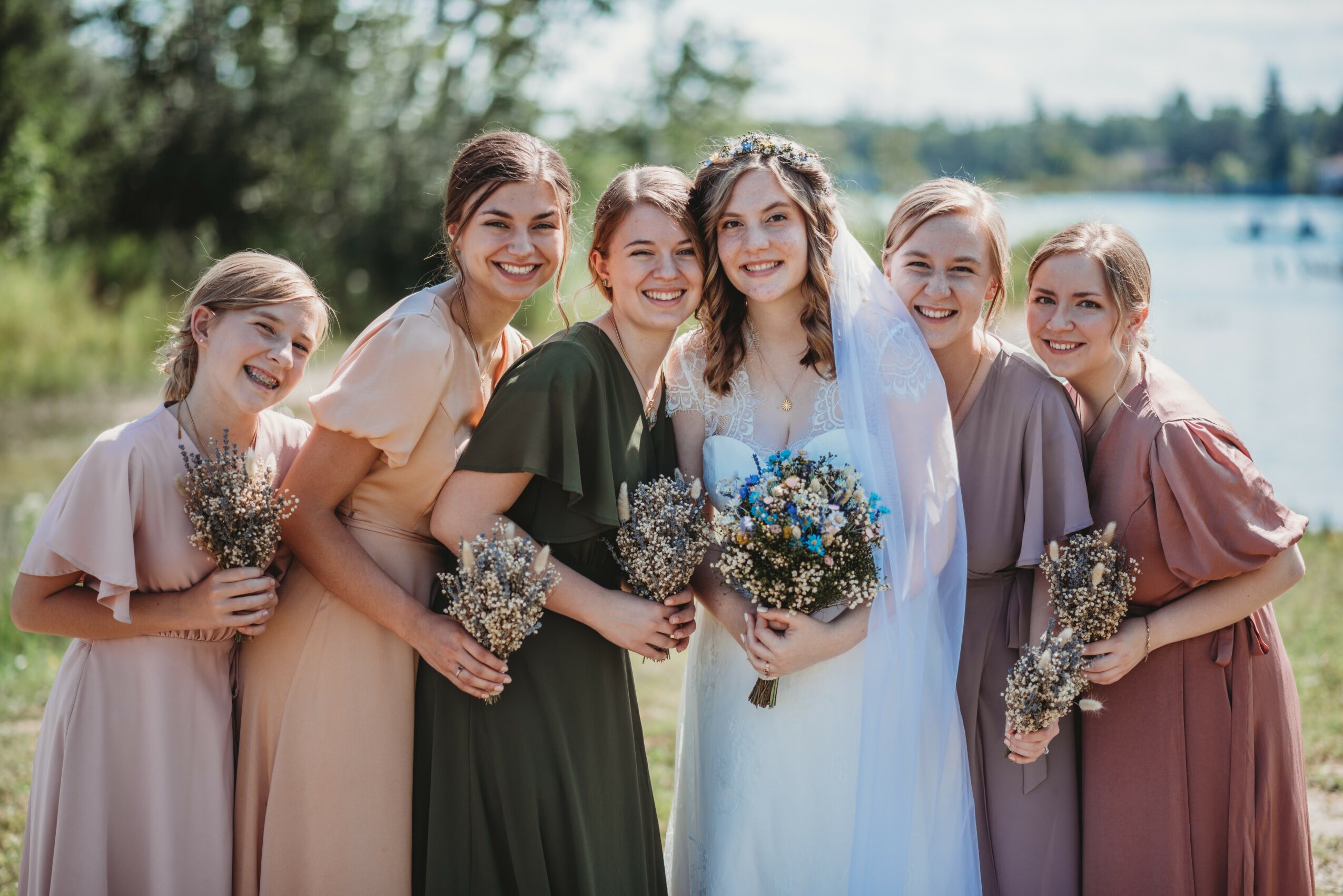 Group of six women outdoors near water, smiling, holding bouquets, dressed in colorful dresses, sunny day.