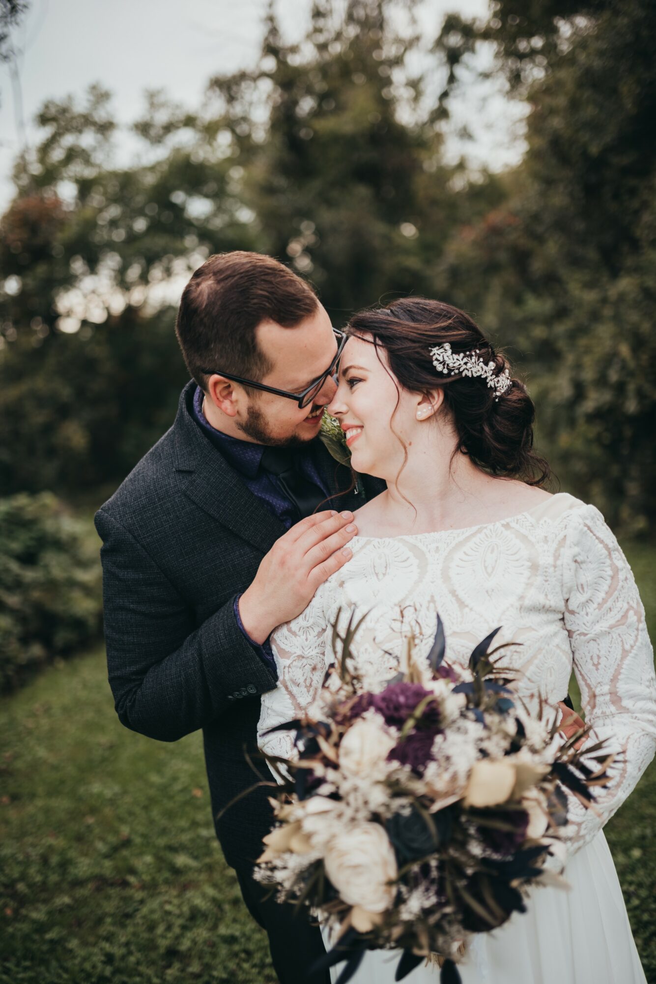 A couple in wedding attire smiling and looking at each other outdoors, with a bouquet of flowers in front.