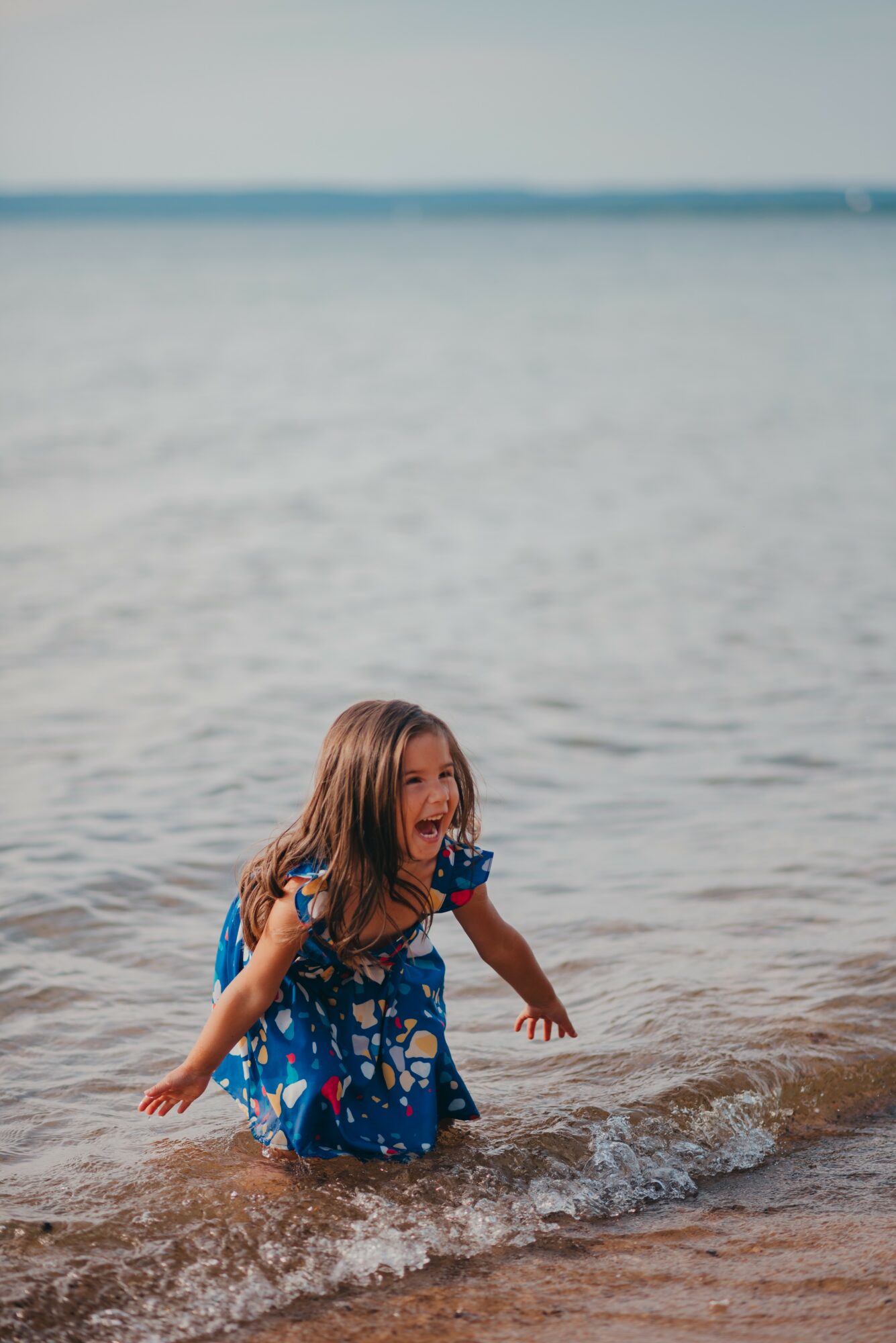 Child in a blue dress playing in shallow water at the beach, smiling and splashing.