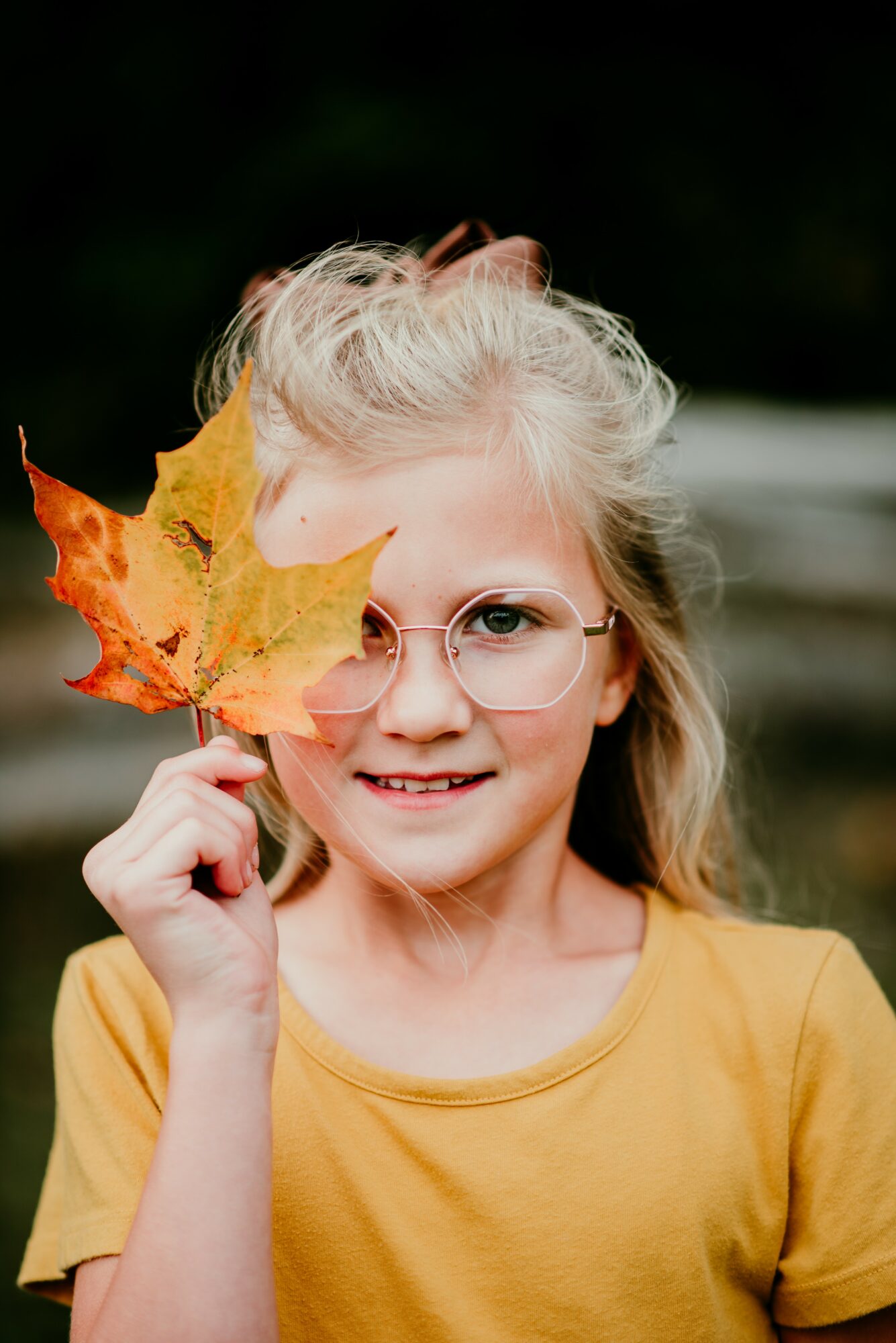 Girl with glasses holding a yellow leaf in front of her face, outdoors, wearing a yellow shirt.