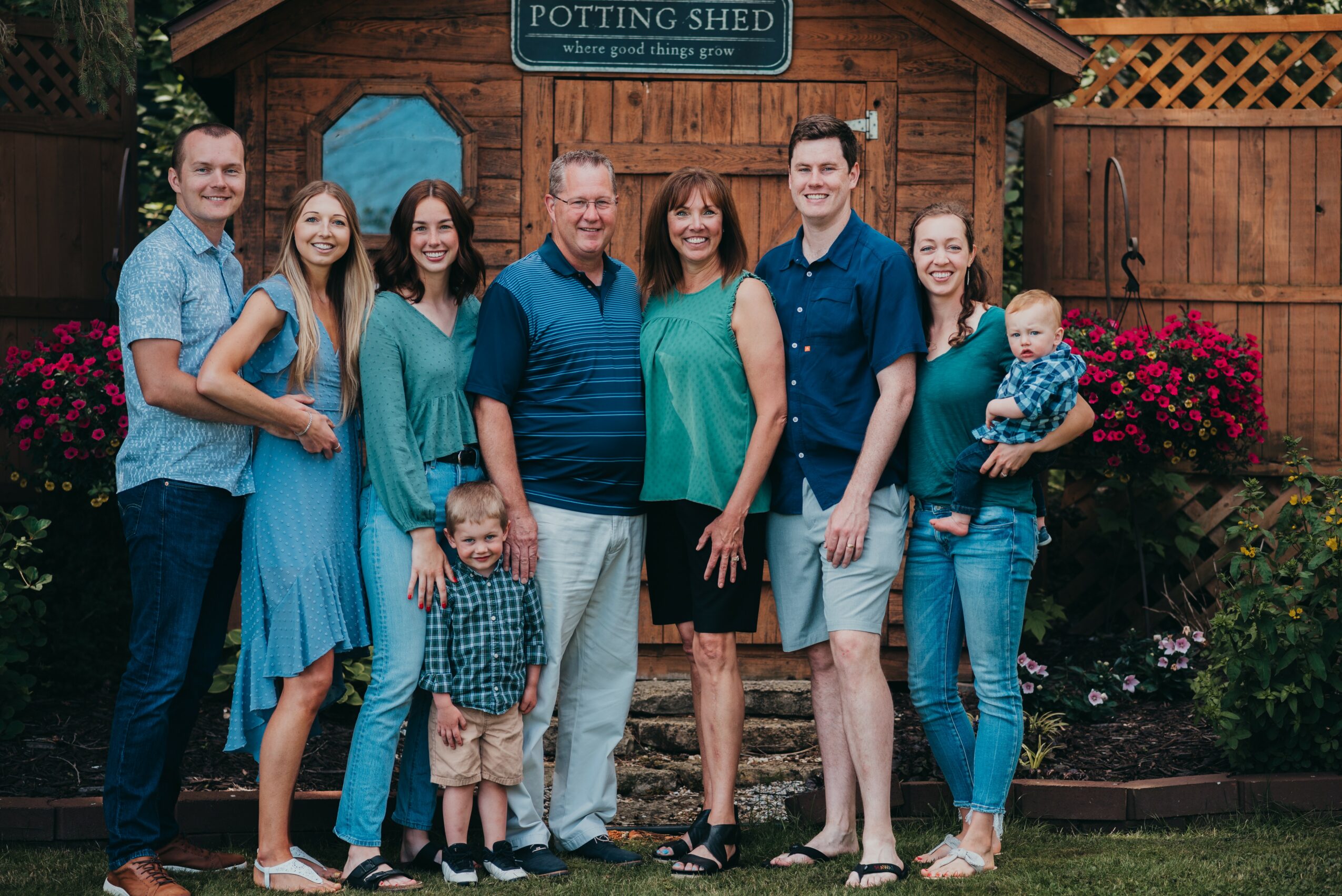 Group of nine people, including children, standing outdoors in front of a wooden shed with a sign, smiling.