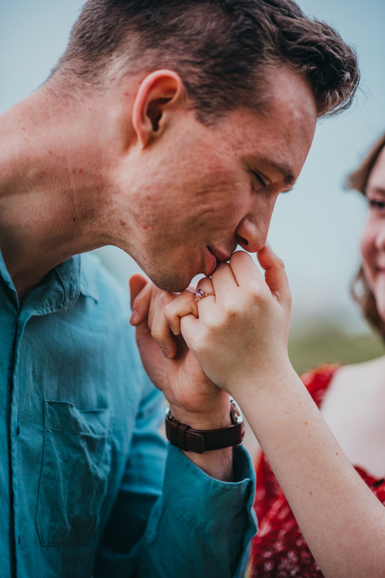 A man and woman hold hands close to their faces, with the man gently kissing the woman's hand outdoors.