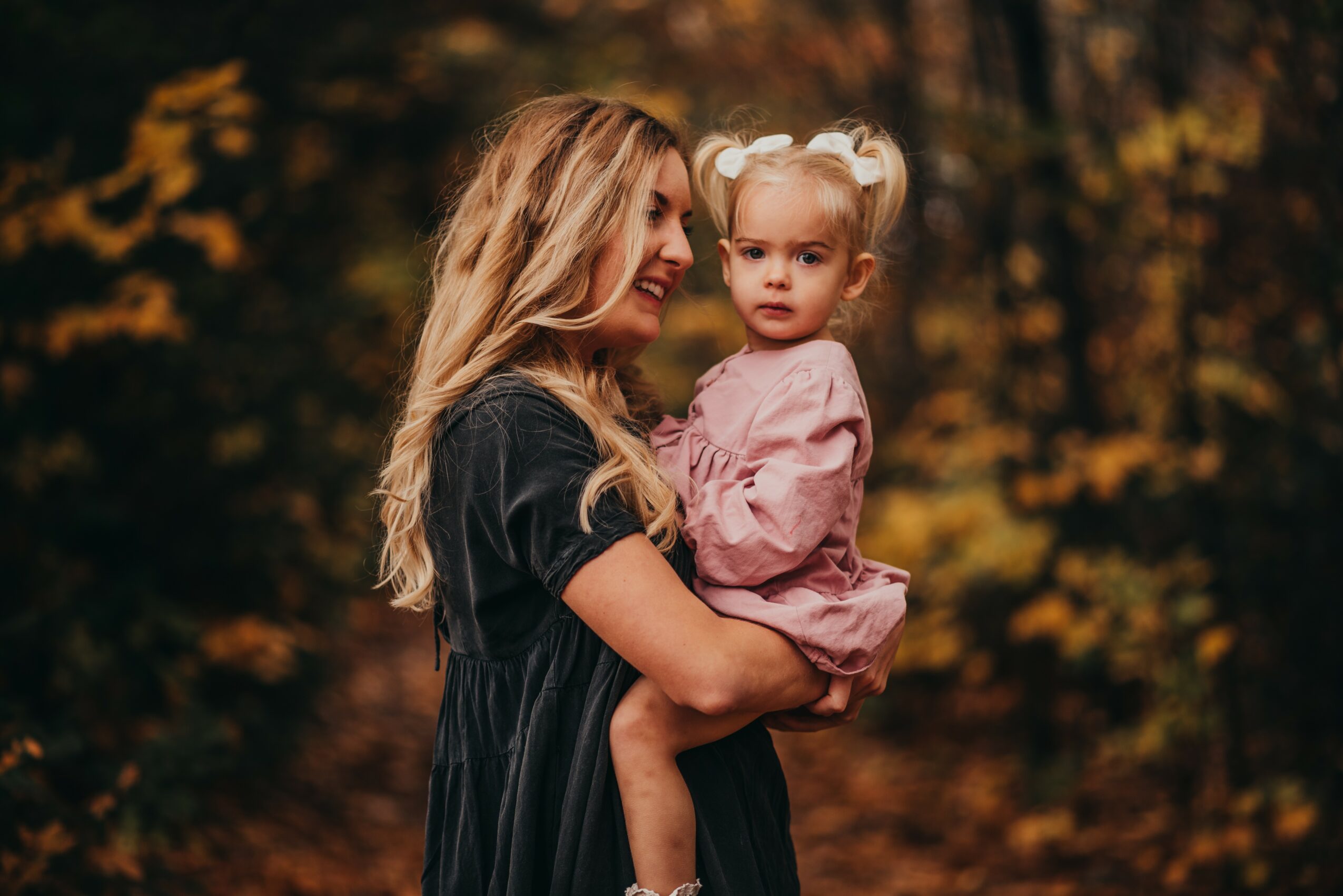 Woman holding a young girl outdoors with autumn foliage in background.