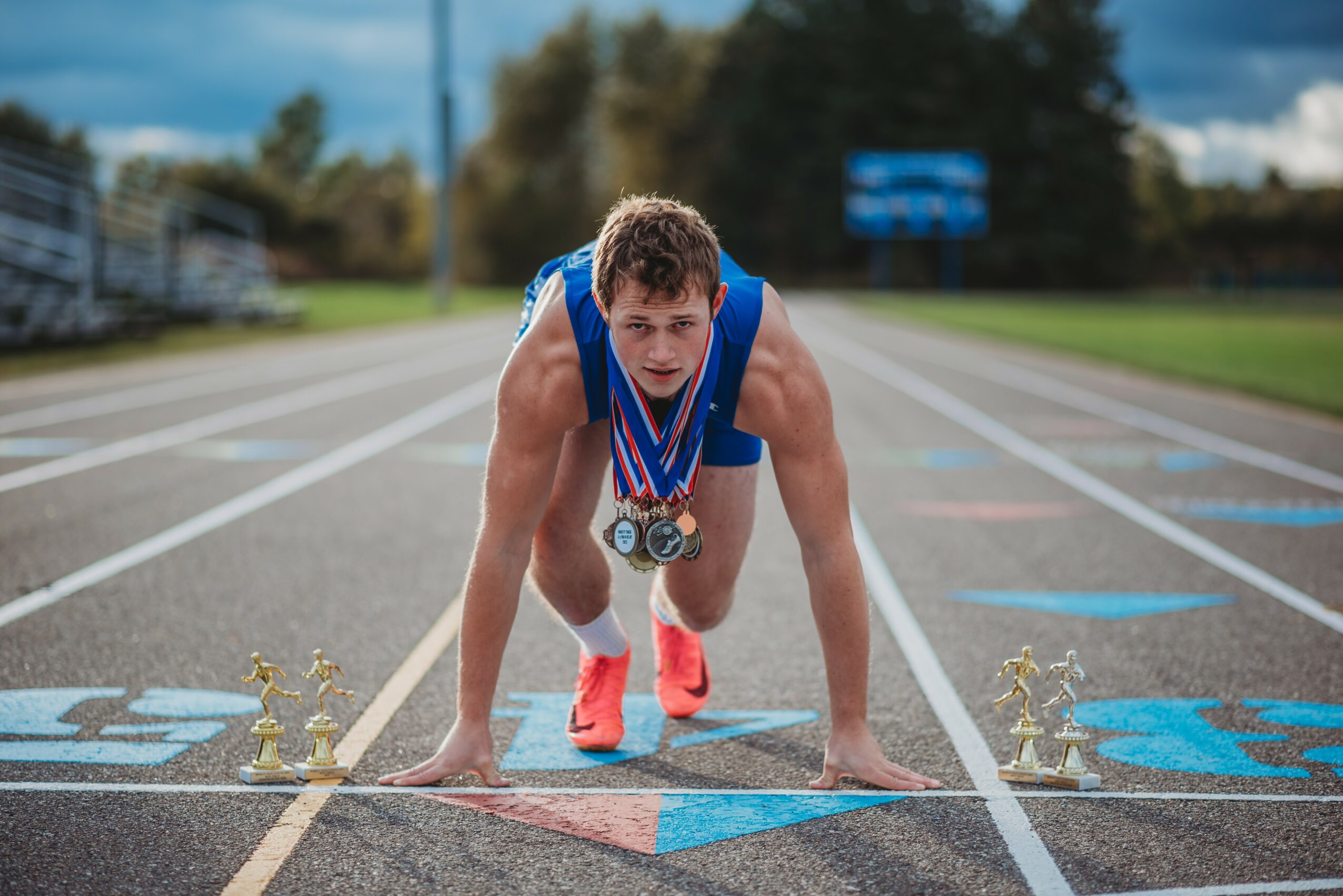 Male athlete in starting position on a track, wearing medals around neck, with trophies nearby, outdoors on a sunny day.