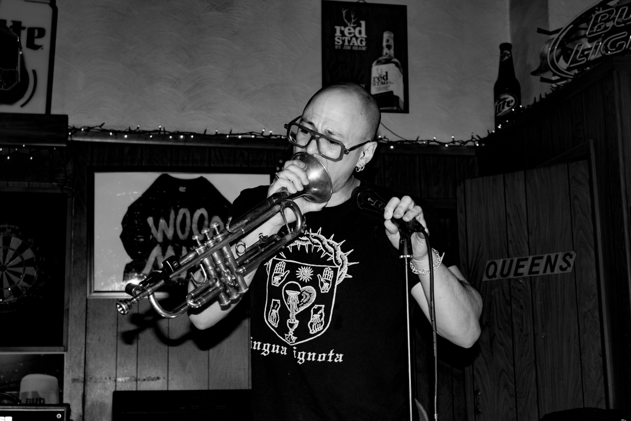 Person with glasses singing into a microphone in a bar, holding a saxophone, with posters and signs in the background.