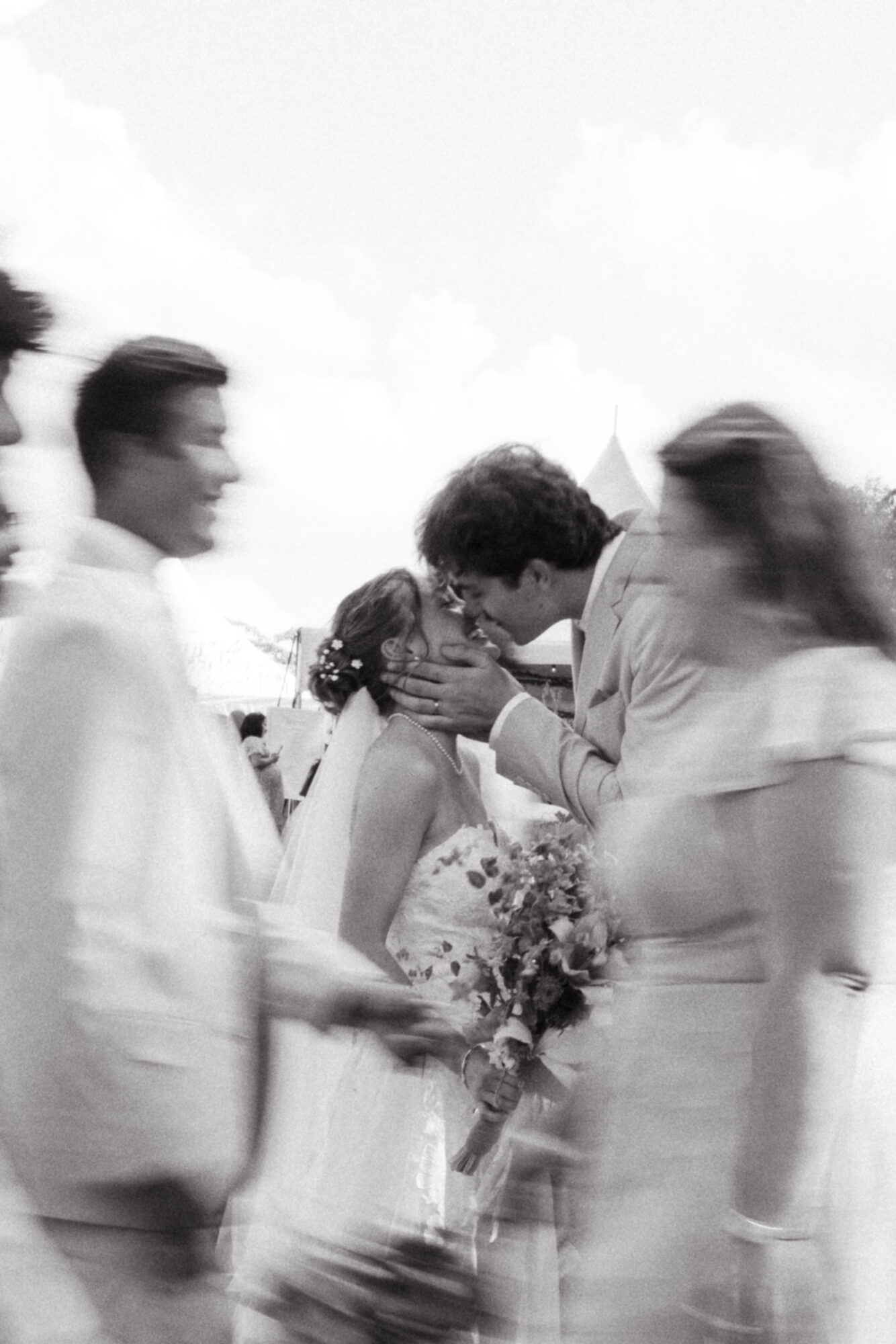 People gather outdoors, with one person kissing another on the cheek, holding a bouquet of flowers, in black and white.
