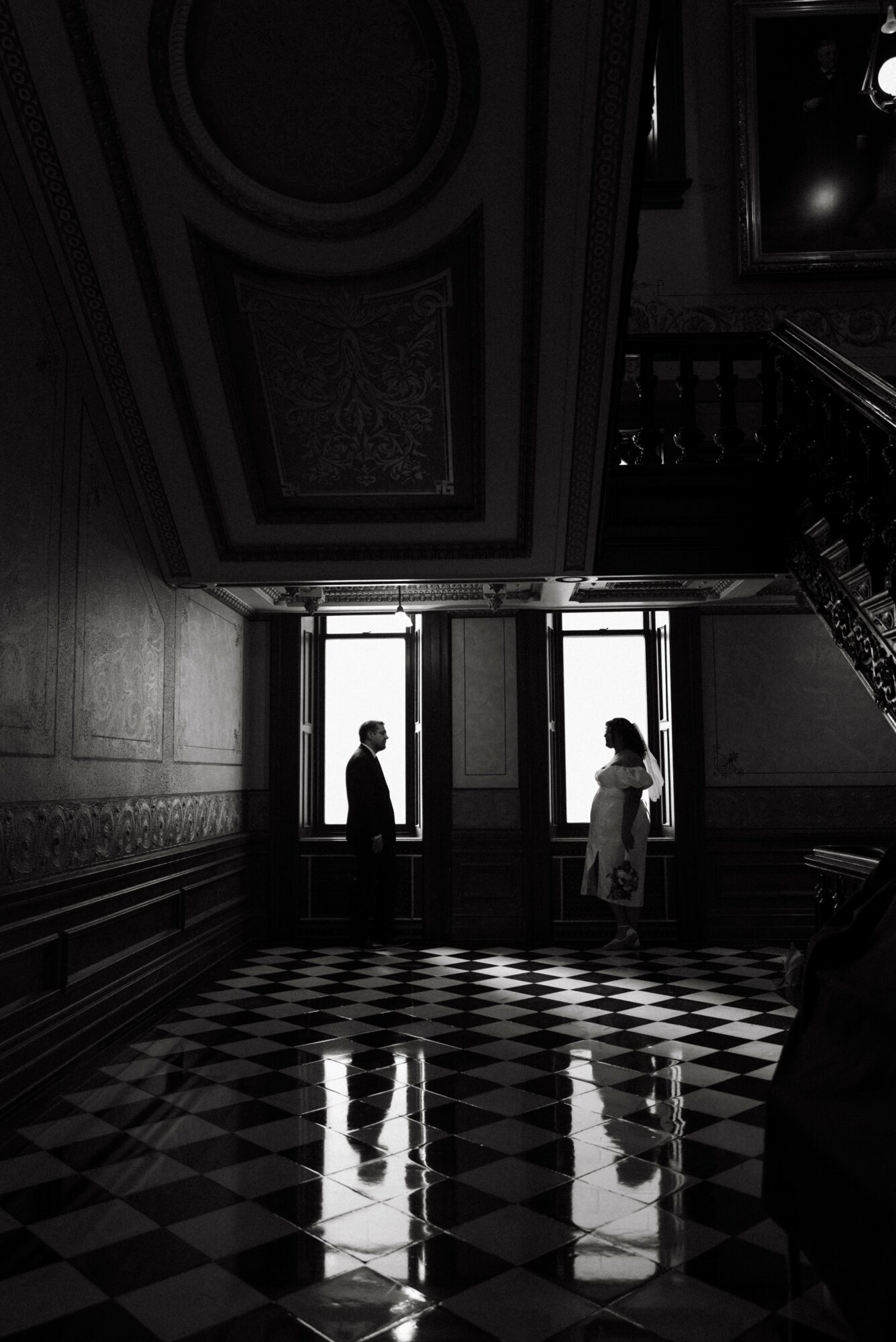 Two people stand facing each other in front of large windows, with reflections on a checkered floor, inside a grand building.