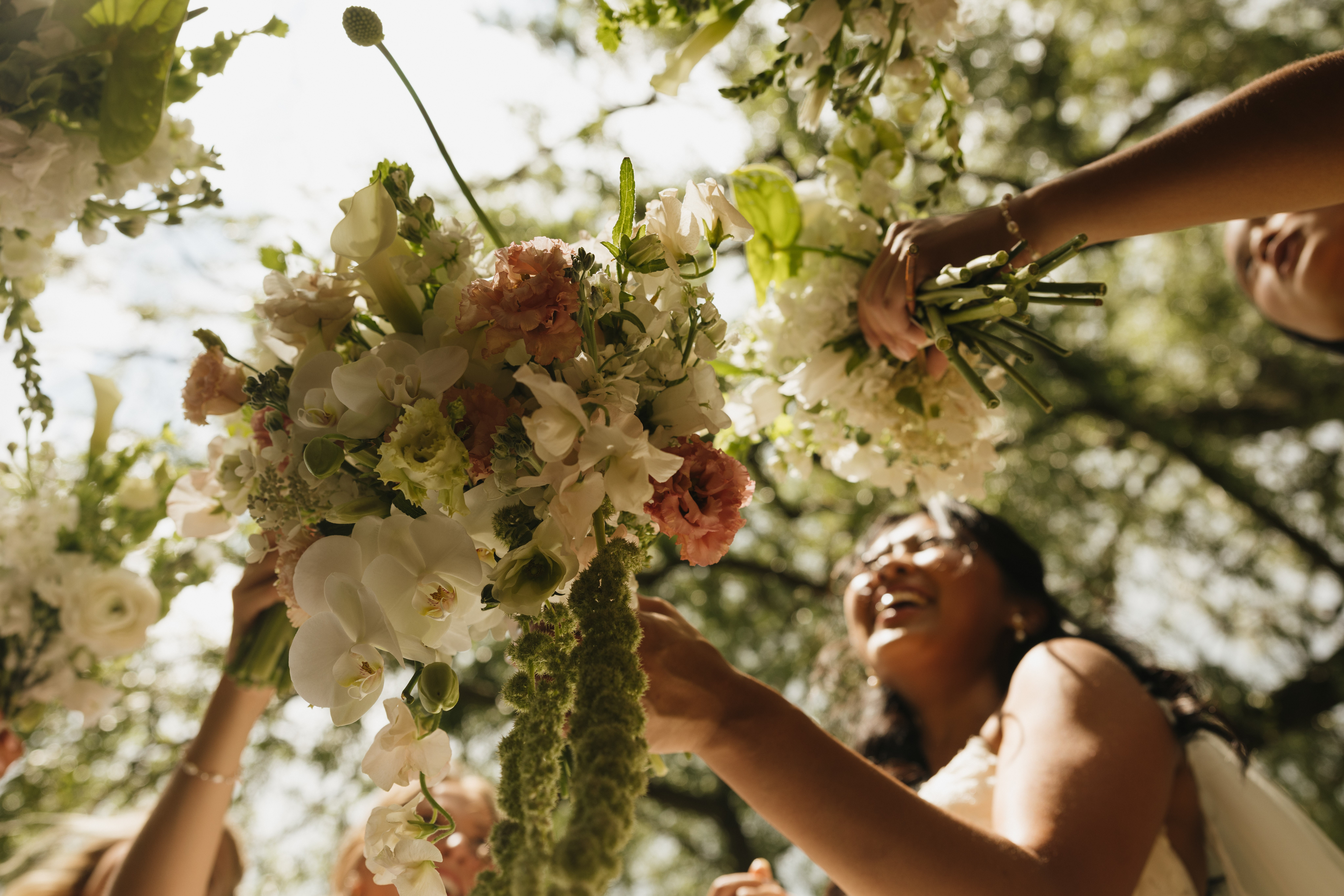People holding bouquets of flowers outdoors, sunlight filtering through trees, smiling and reaching upward.