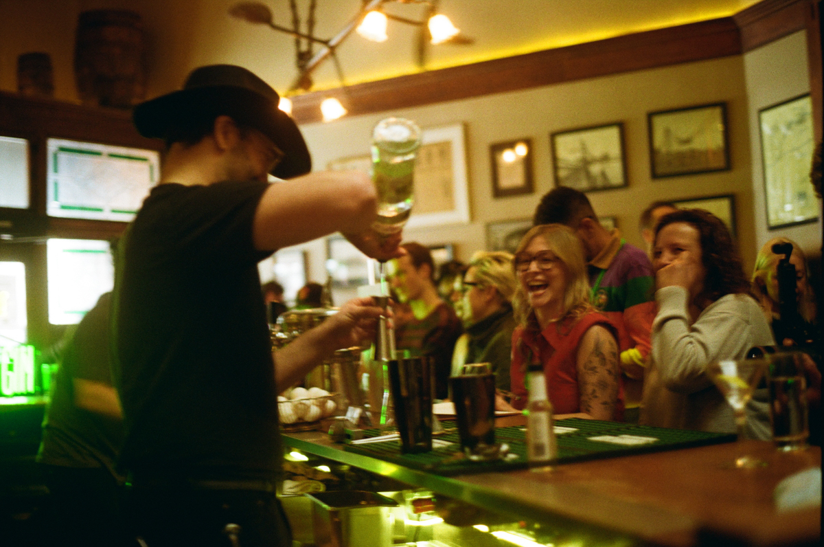 Bartender pouring drink for smiling group at bar, warm lighting, framed pictures on wall, lively atmosphere.