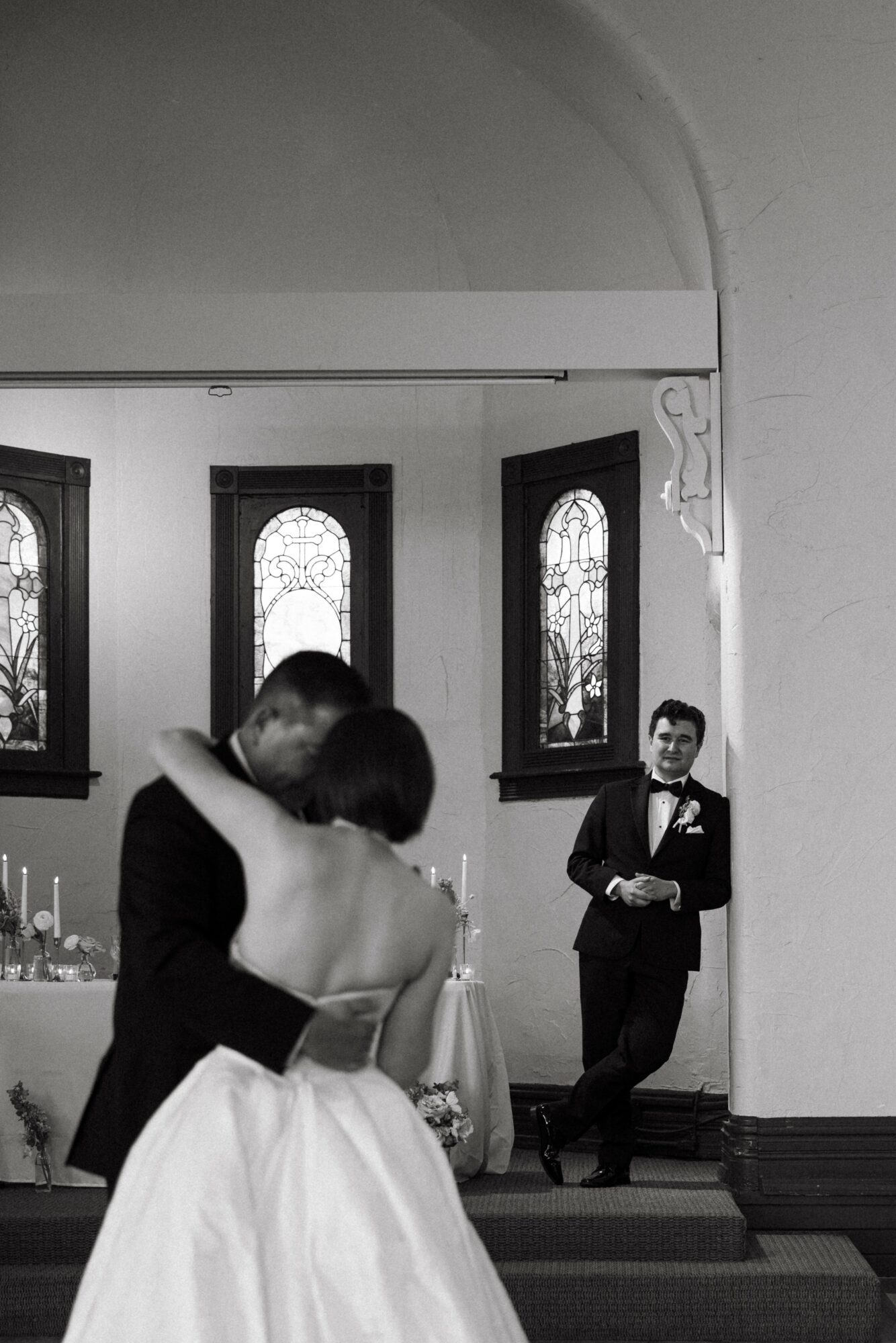 A couple dancing in a church with stained glass windows, a man in a tuxedo watching, and a woman in a strapless dress.