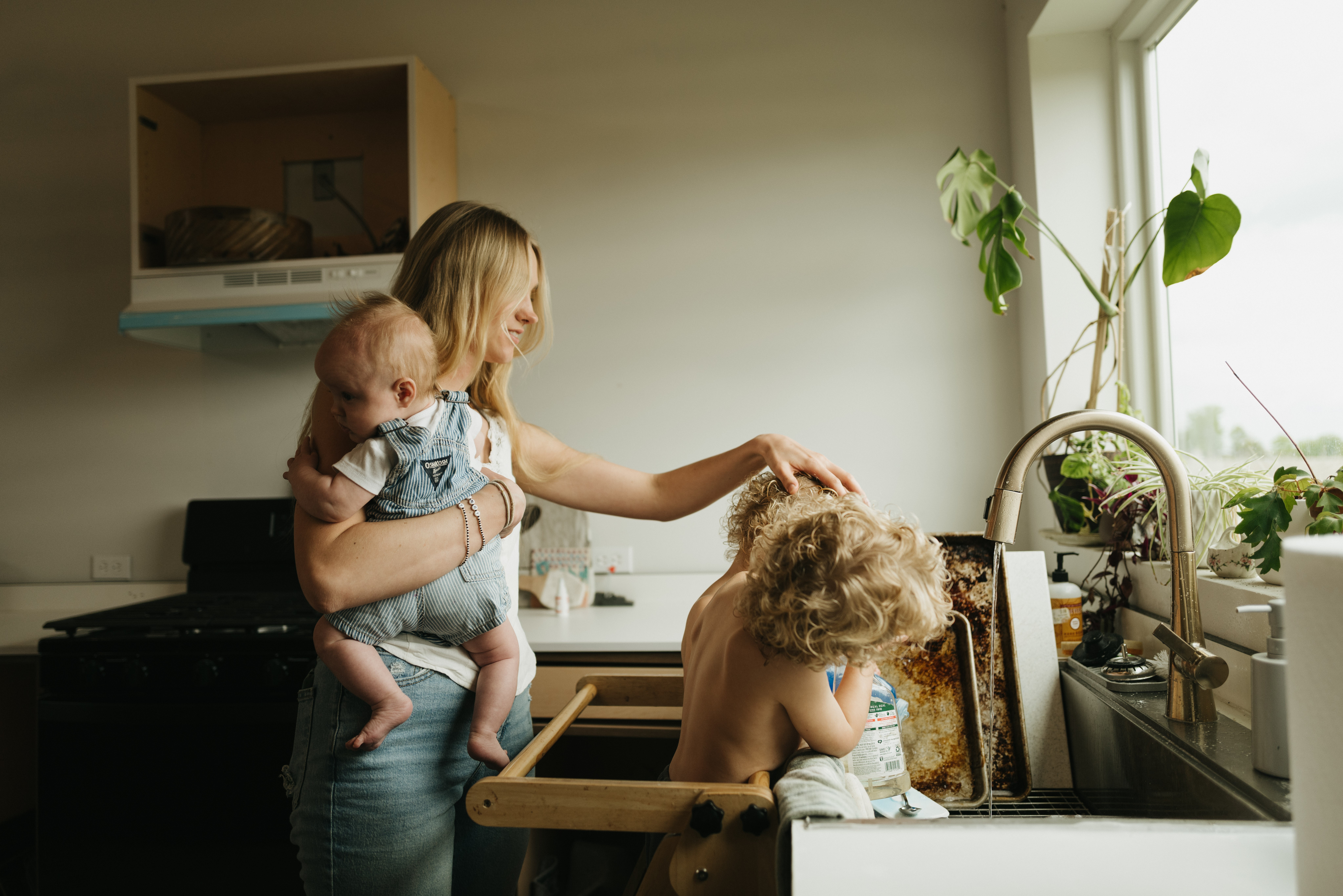 Woman holding a baby in a kitchen, with a child leaning over the sink, sunlight coming through a window, plants nearby.