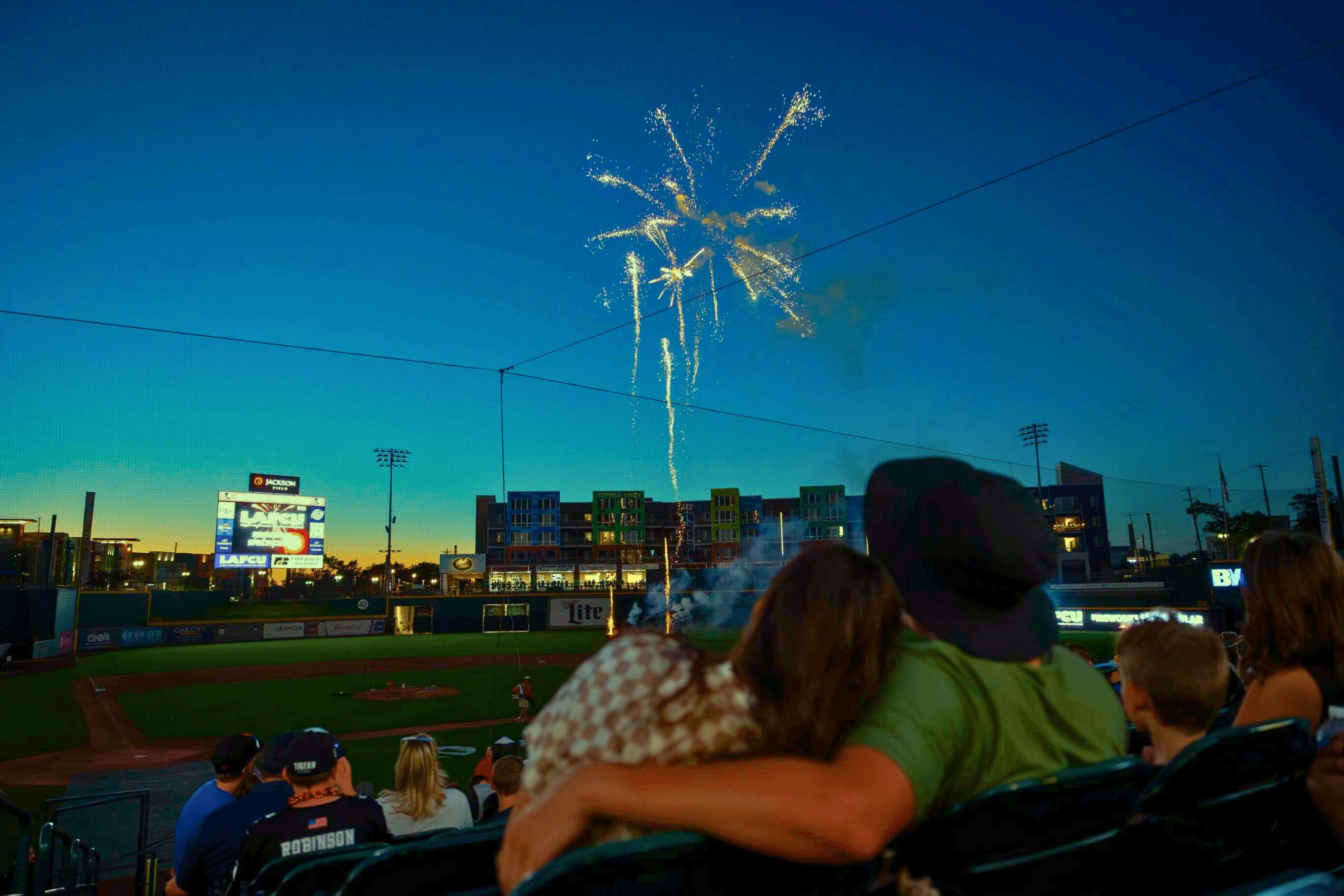 People watching fireworks in a stadium at dusk, city buildings in the background, some with illuminated signs.