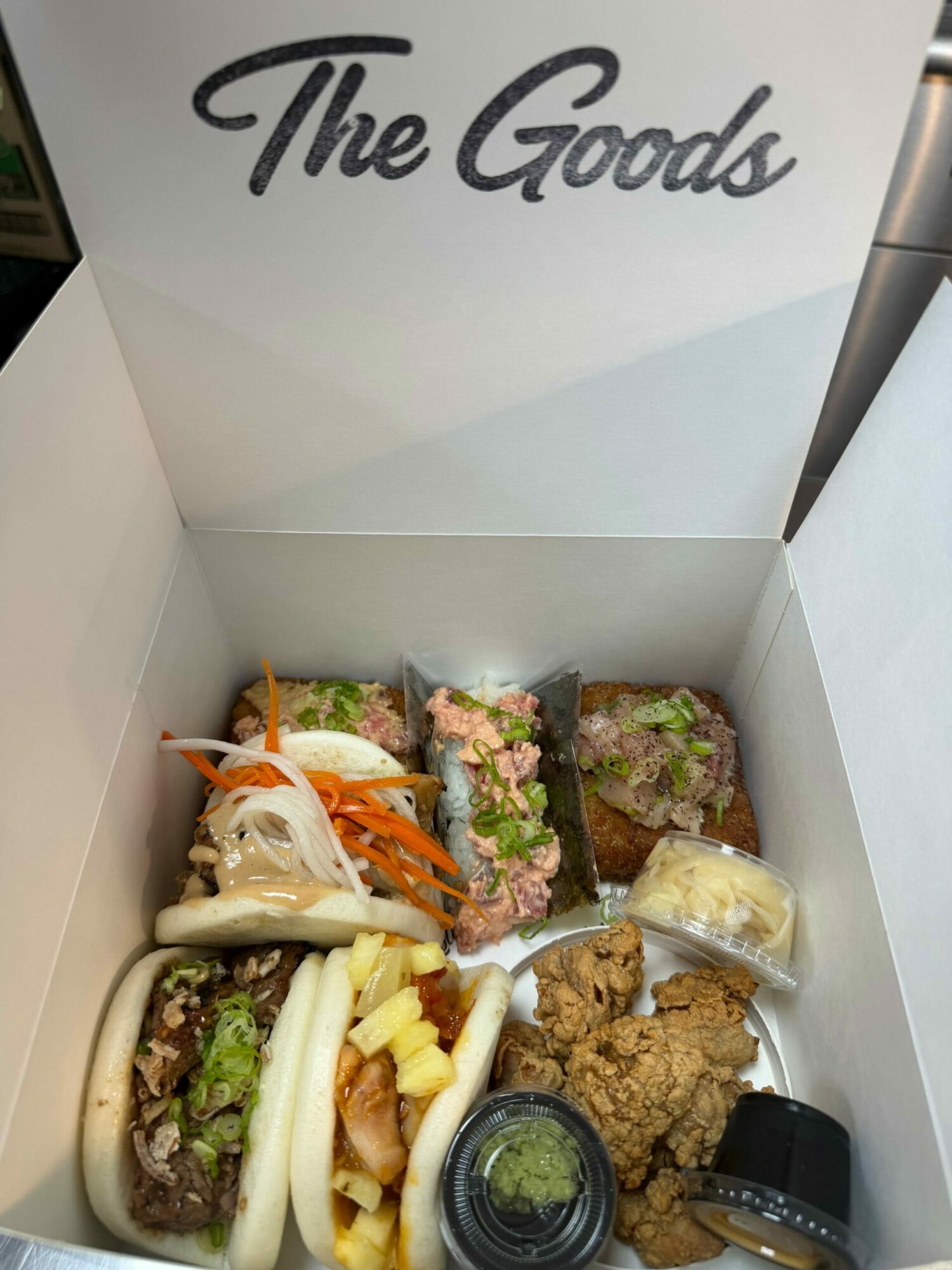 Open box with three sandwiches, potato chips, and small containers of condiments inside. The box has 'The Good' written on the inside lid.