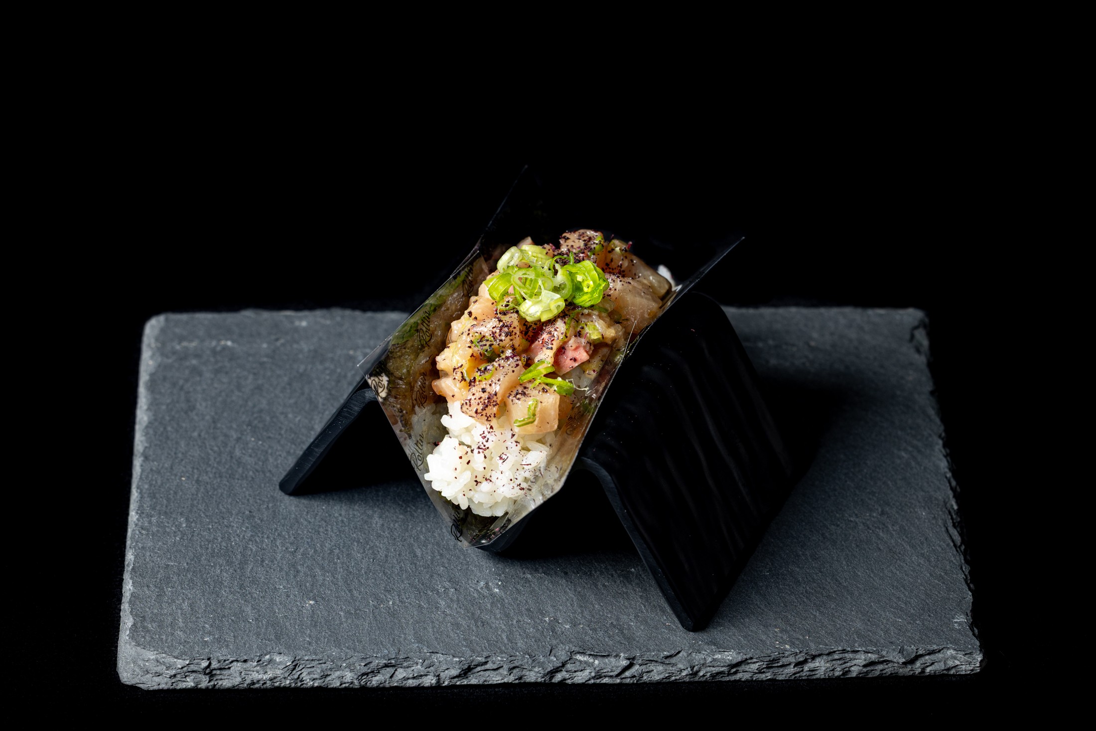 Sushi roll with rice, fish, and green garnish on a black slate plate against a black background.